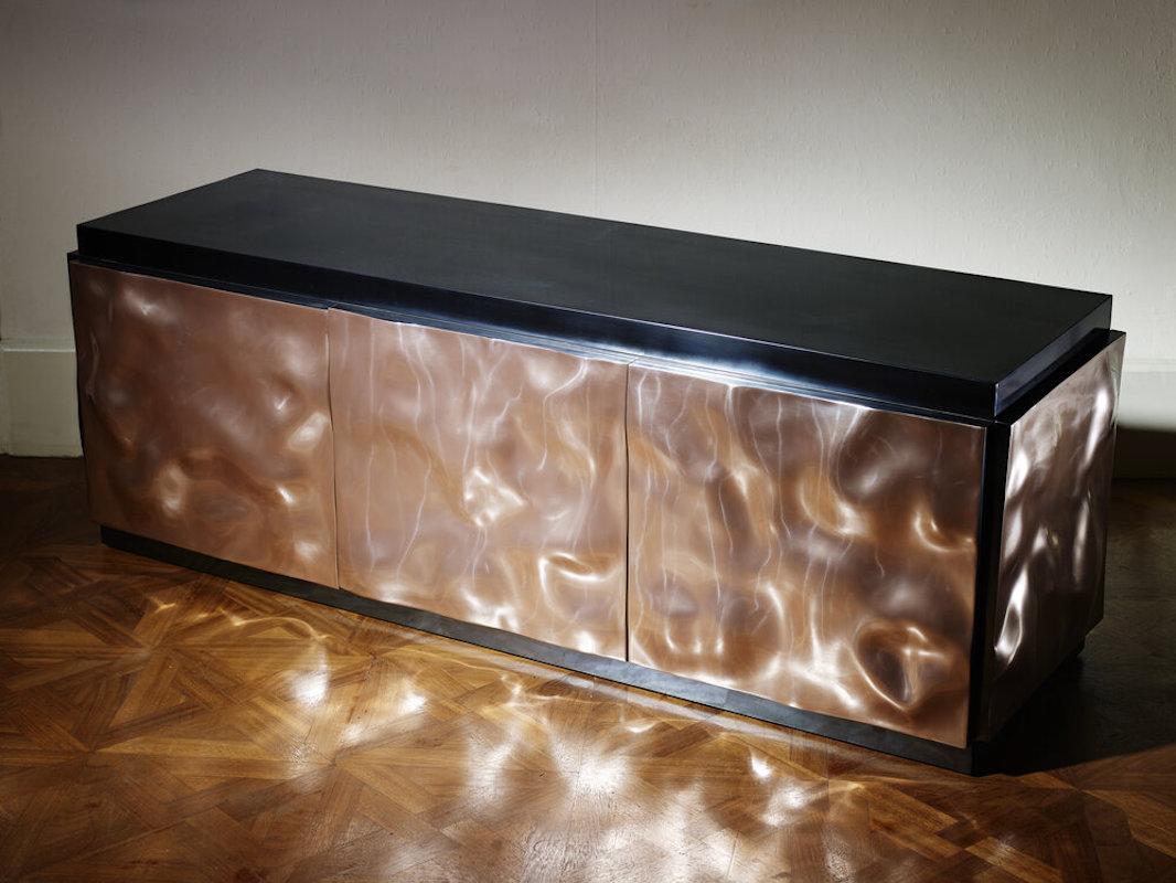 Bumper is a sideboard with three doors. By combining two materials (the pink copper of the doors and the deep black of the oxidized zinc top with its bluish glints) and two textures (the bumpy appearance of the doors and the smoothness of the top),