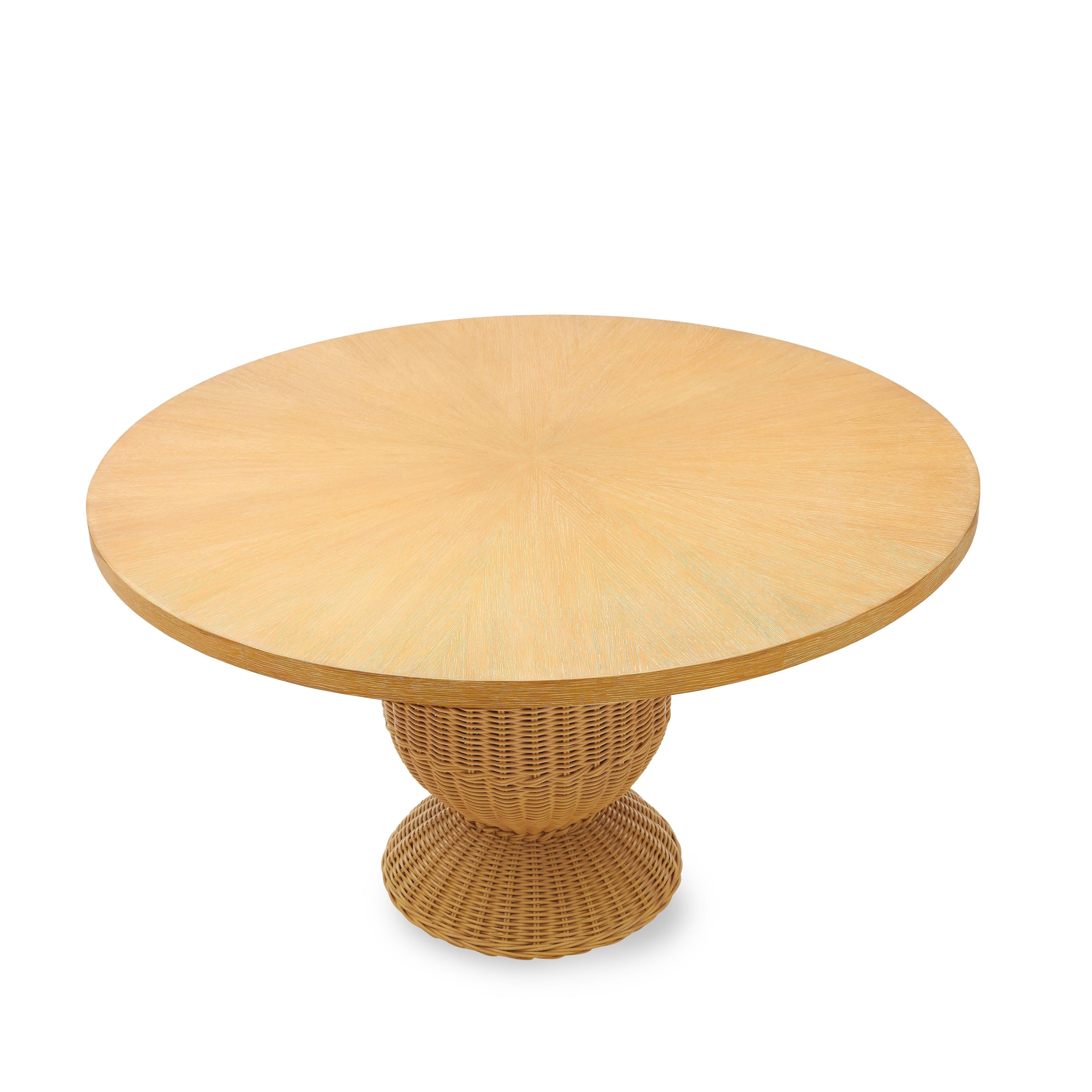Our Braided Urn Dining Table features a gracefully curved rattan base, woven in an urn-like silhouette with intricate braided details. A round, grey-washed oak top adds a refined contrast, while a hidden metal support ensures durability. A warm,
