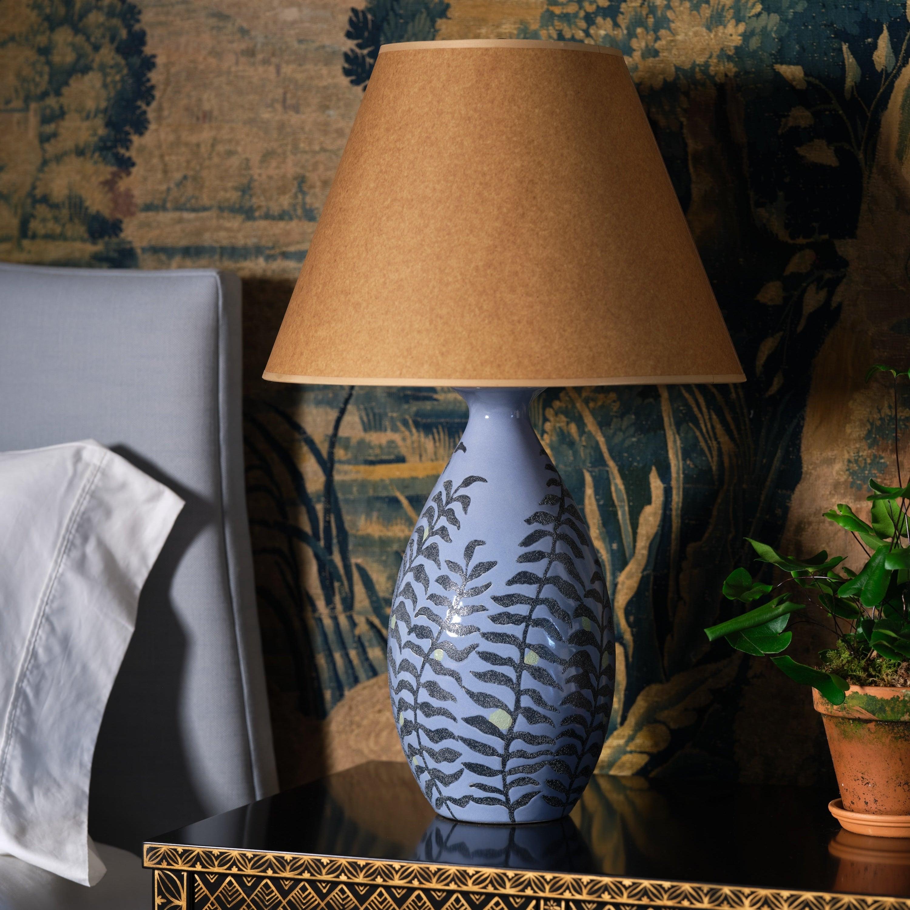 A delicate fern motif graces this lamp, its fronds gracefully unfurling along the vase, a nod to nature’s quiet beauty. The silhouette is svelte and refined, designed to complement any space with understated elegance. The calming blue glaze, coupled