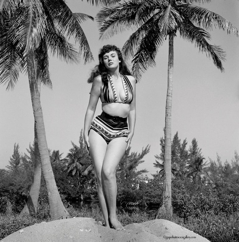 Bunny Yeager, the pre-eminent female pin-up photographer of the 20th century took this pin-up self-portrait in 1953. The artist and model showcases her draw on both sides of the camera by posing atop a sandy mound, bookended by two palm trees, and