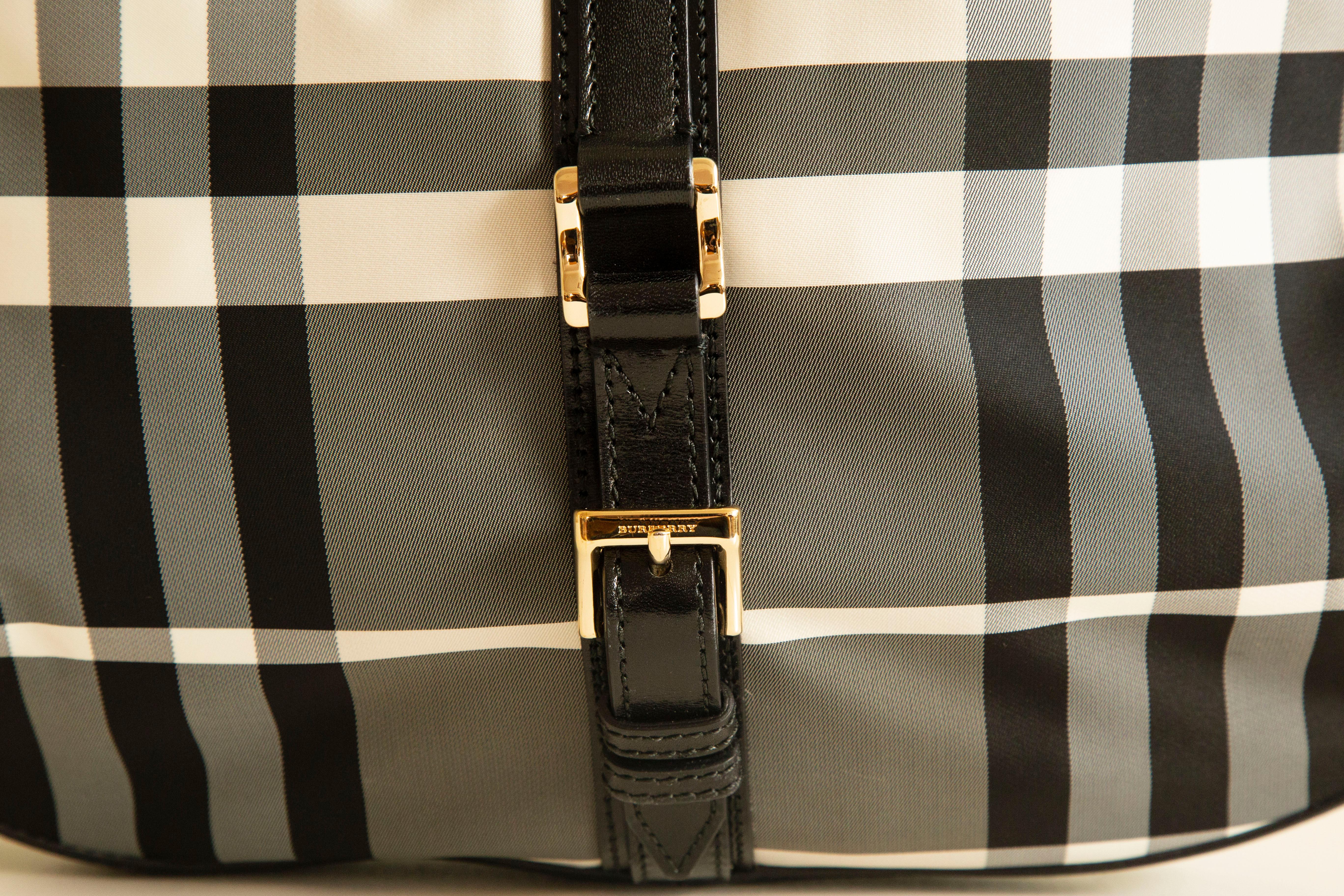 Sac à bandoulière/hobo bag spacieux de Burberry en nylon à motif Nova Check noir/gris/blanc. Le sac est orné d'une bordure en cuir noir et de ferrures dorées. L'intérieur se compose d'un compartiment principal et de trois poches latérales, dont