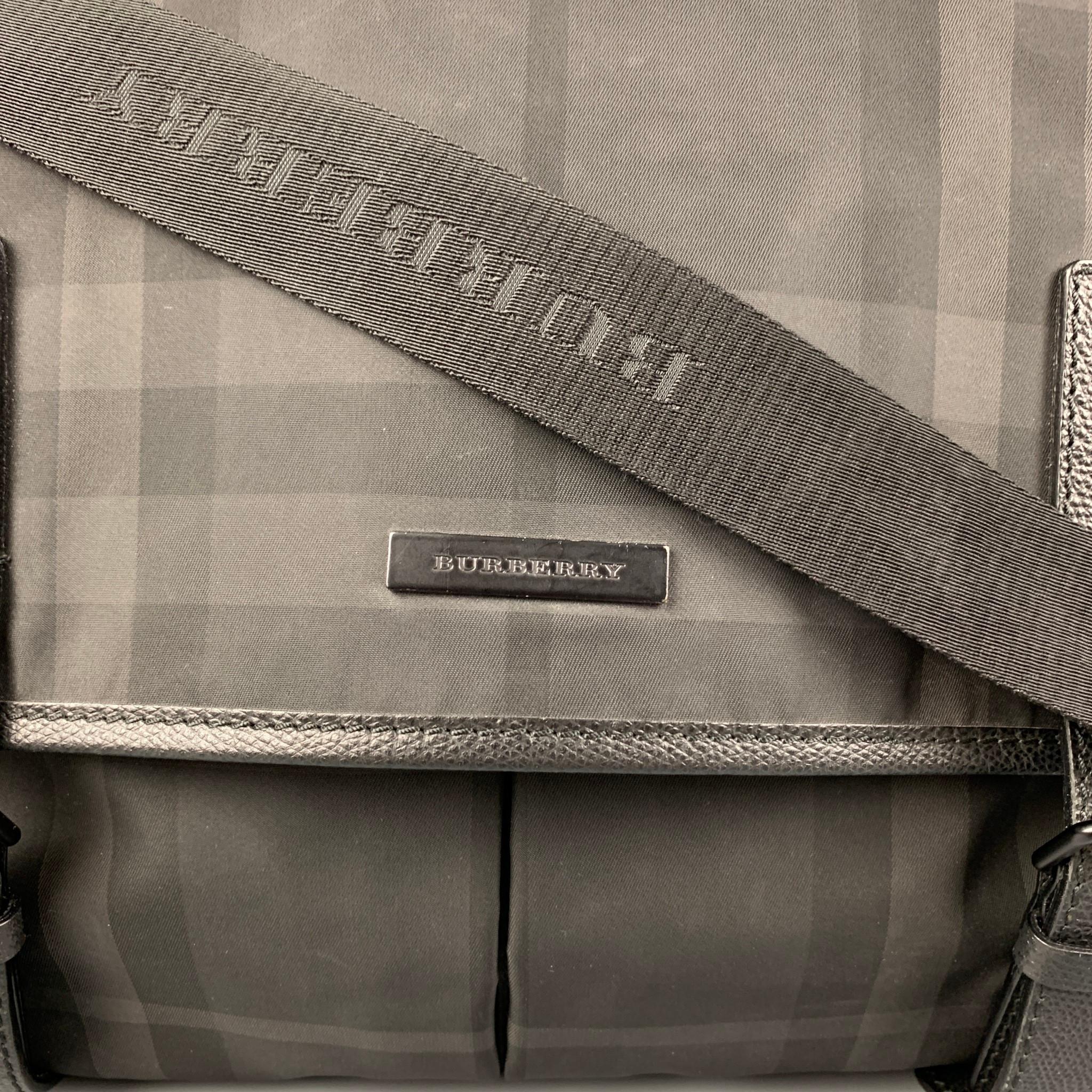 BURBERRY bag comes in a black & grey plaid nylon featuring a messenger style, adjustable cross body strap, leather trim, back pocket, inner pockets, and a snap button closure. Made in Italy. 

Very Good Pre-Owned Condition.
Marked: