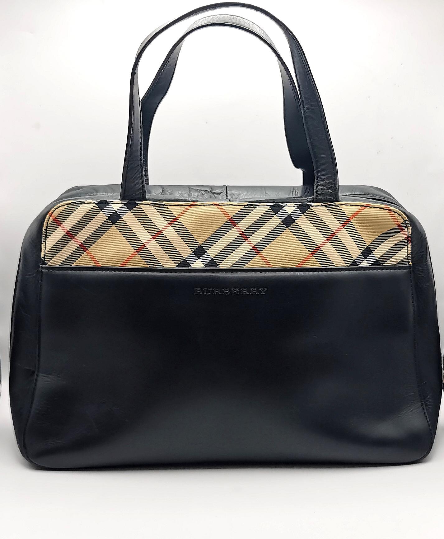 Un elegante bolso Burberry estilo Boston en piel negra.

Se trata de un bolso de tamaño medio con asas superiores de piel y el característico ribete de cuadros Burberry en la parte superior delantera del bolso.

Tiene un cierre de cremallera con una