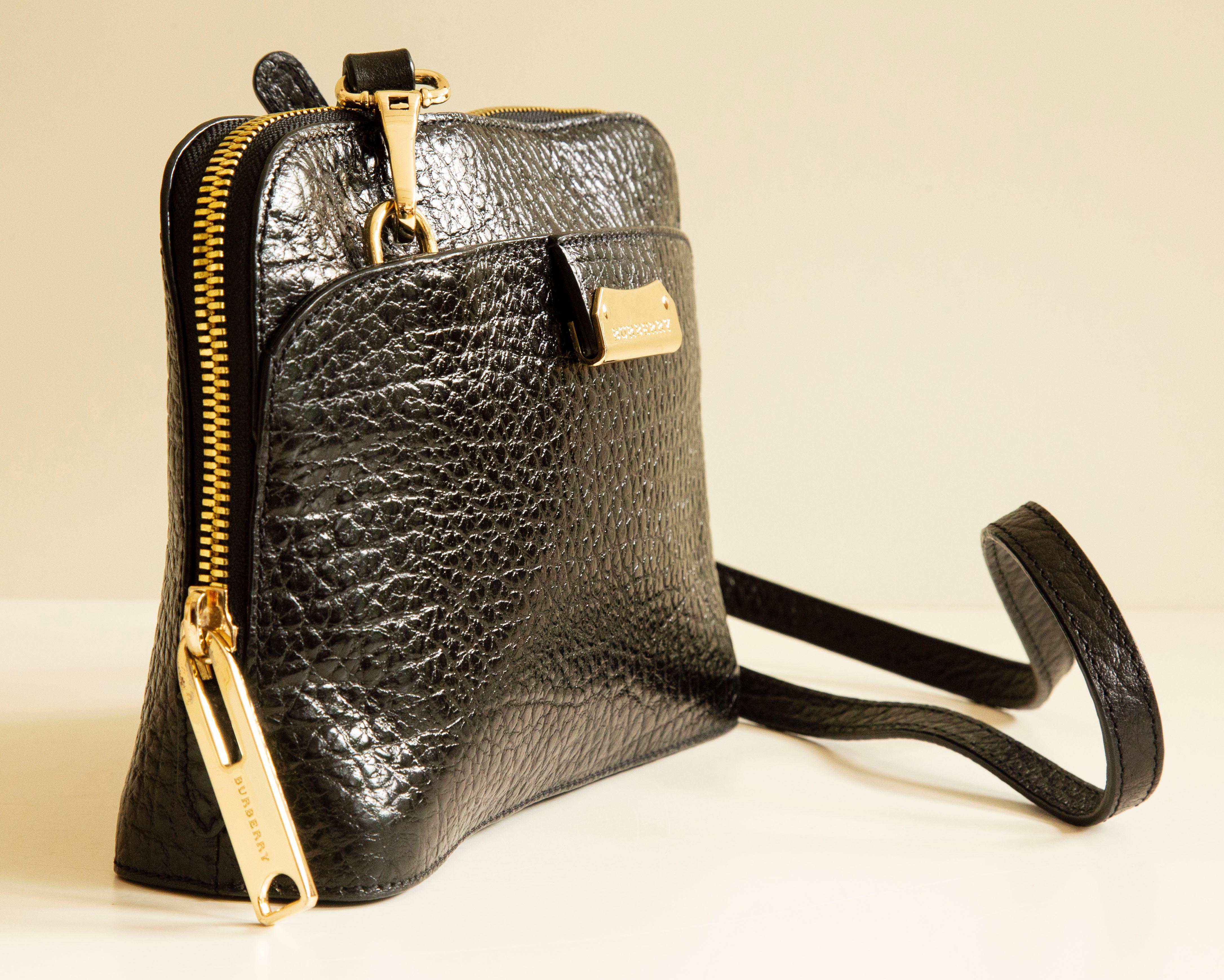 A Burberry shoulder bag/crossbody bag  made of black shiny and textured leather with gold toned hardware. The interior is lined with black fabric and it features a major compartment and two side pockets of which one has a zipper. The shoulder strap
