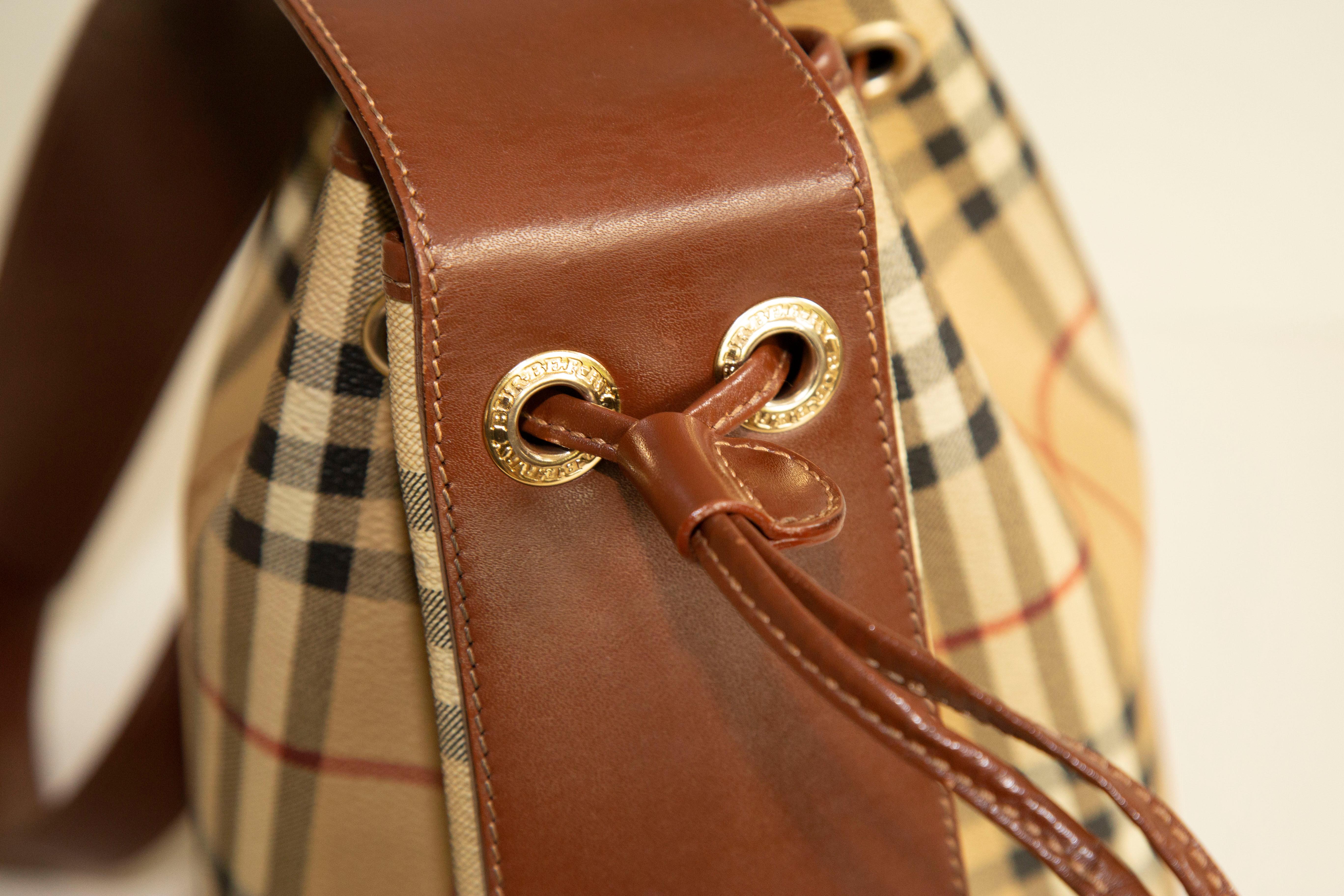 A Burberry Bucket bag. The bag features a classic check pattern exterior, brown leather trim, and gold-toned hardware. The interior is lined with black fabric and there is one side pocket with a zipper. The interior is neat & clean. The exterior,