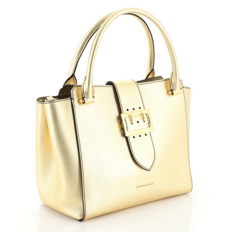 This Burberry Buckle Tote Leather Medium, crafted from gold leather, features dual rolled leather handles, flap tab with buckle detail, protective base studs, and gold-tone hardware. Its magnetic snap button closure opens to a brown house check