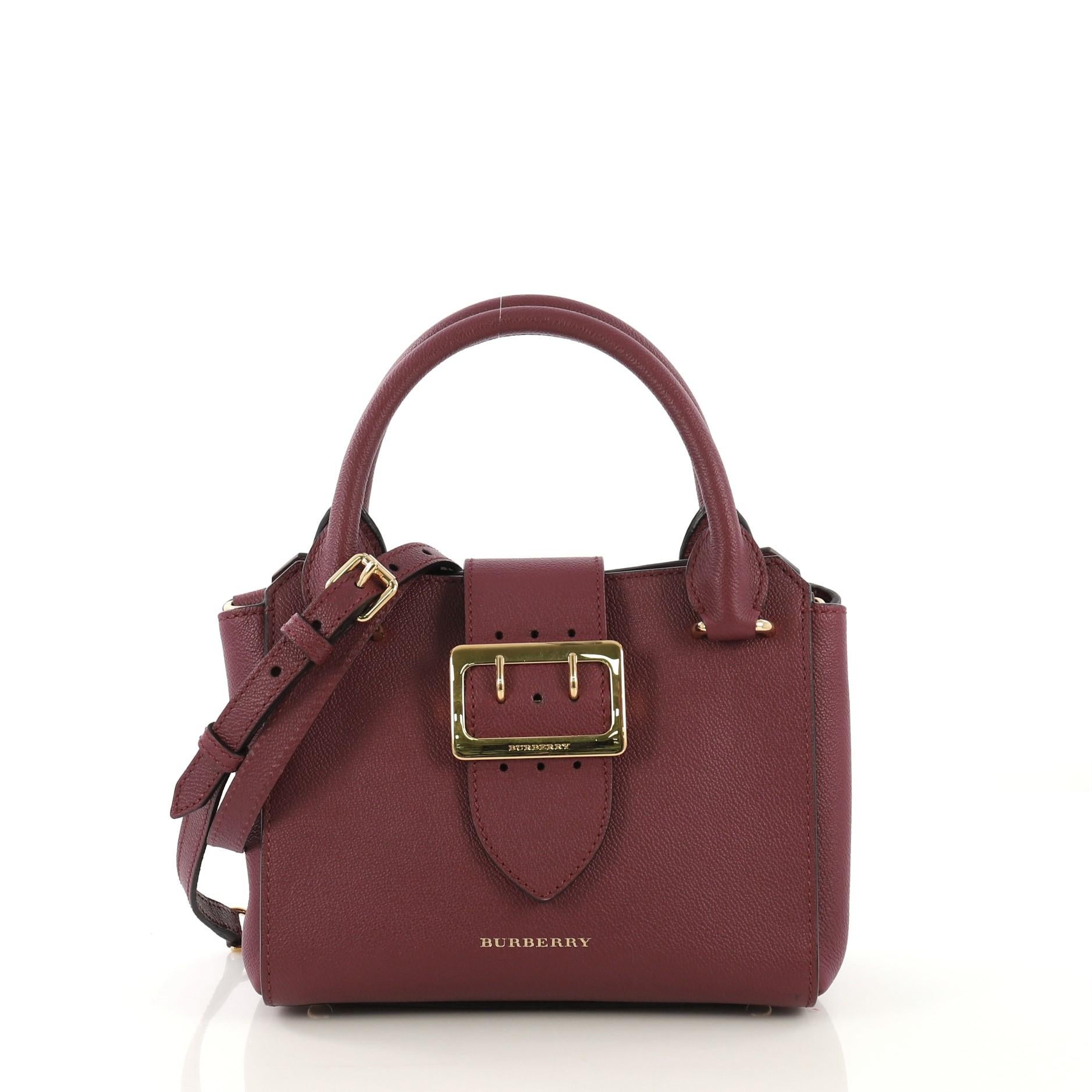 This Burberry Buckle Tote Leather Small, crafted from purple leather, features dual rolled leather handles, flap tab with buckle detail, and gold-tone hardware. Its magnetic snap button closure opens to a house check print fabric interior with zip