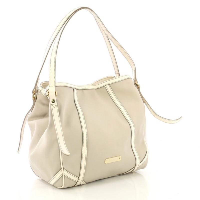 This Burberry Canterbury Tote Trench Canvas Medium, crafted in gray trench canvas, features leather shoulder straps, protective base studs, leather trim, and gold-tone hardware. Its magnetic snap closure opens to a brown fabric interior with zip and