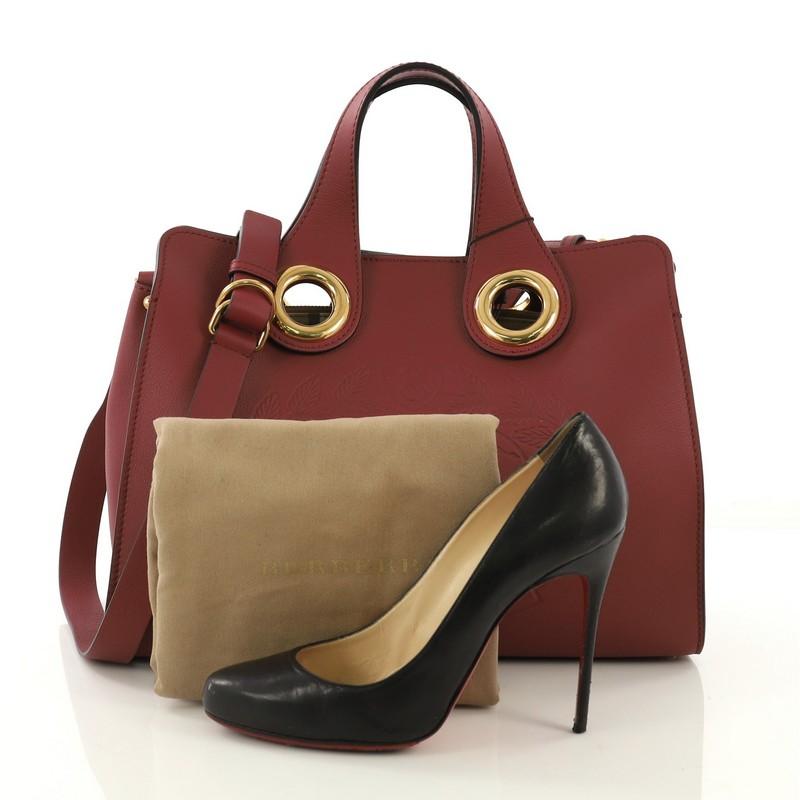 This Burberry Crest Grommet Tote Embossed Leather Large, crafted from burgundy embossed leather, features dual top handles, polished grommets, and gold-tone hardware. Its magnetic snap closure opens to a brown check print and suede interior with