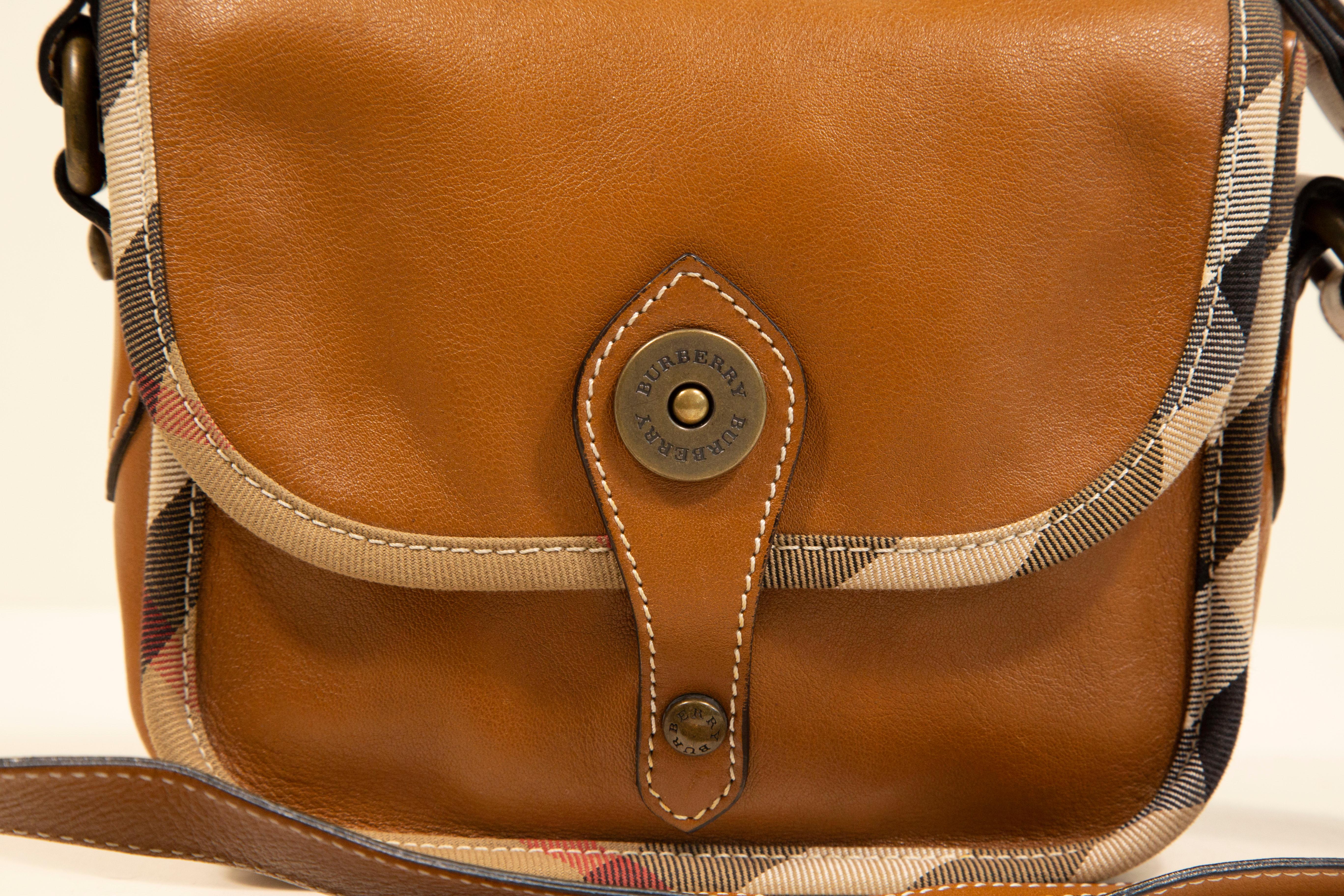 A Burberry crossbody bag made of light brown leather with the Burberry check canvas trim and old brass toned hardware. The interior is lined with brown canvas, and next to the major compartment it features one side pocket with a zipper and one slide
