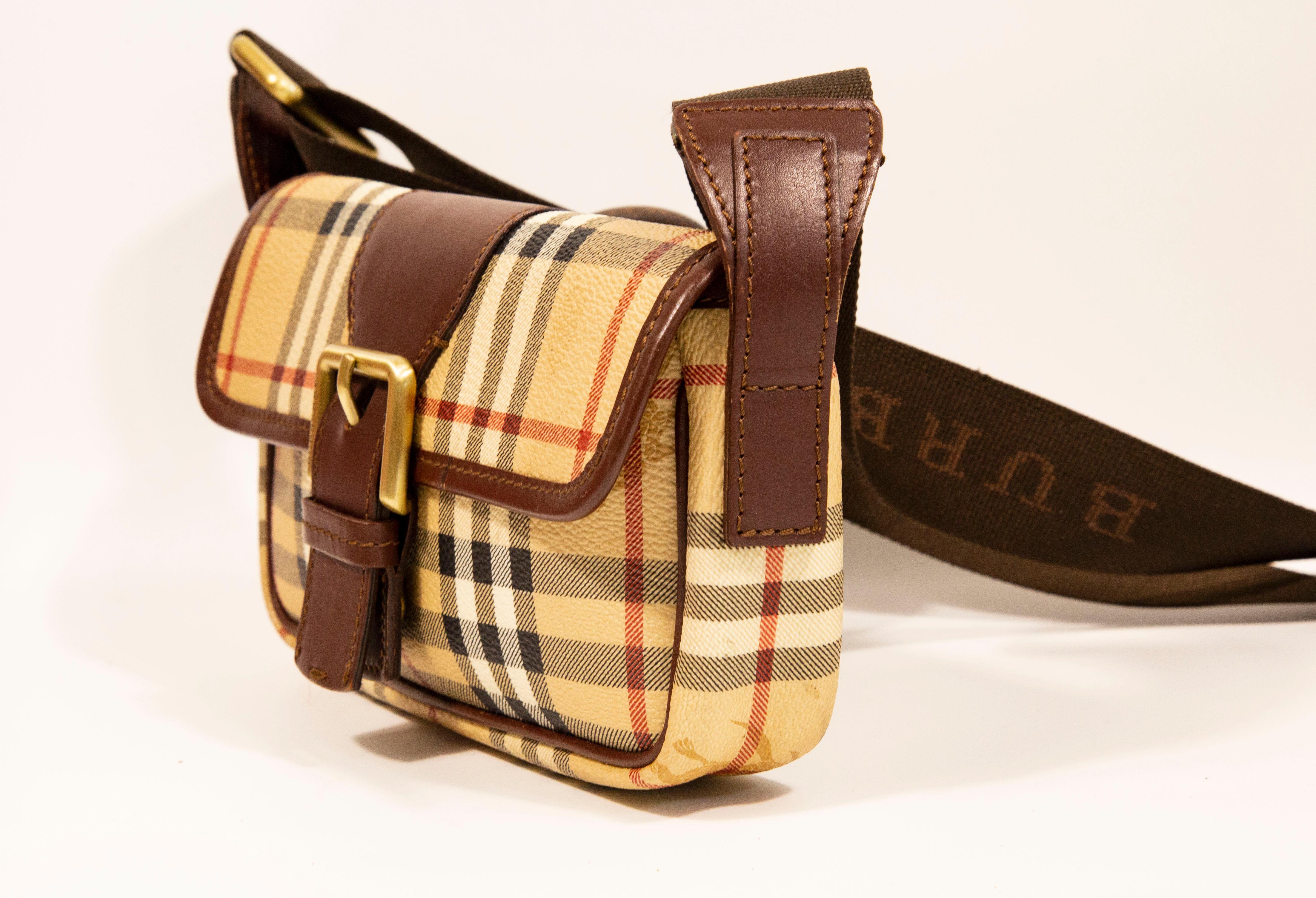 A small Burberry corssbody bag made of vinyl coated canvas with the Burberry classic check, brown leather trim, and brass toned hardware. The bag features a canvas shoulder strap that is in length adjustable max, 114 cm. The interior is lined with