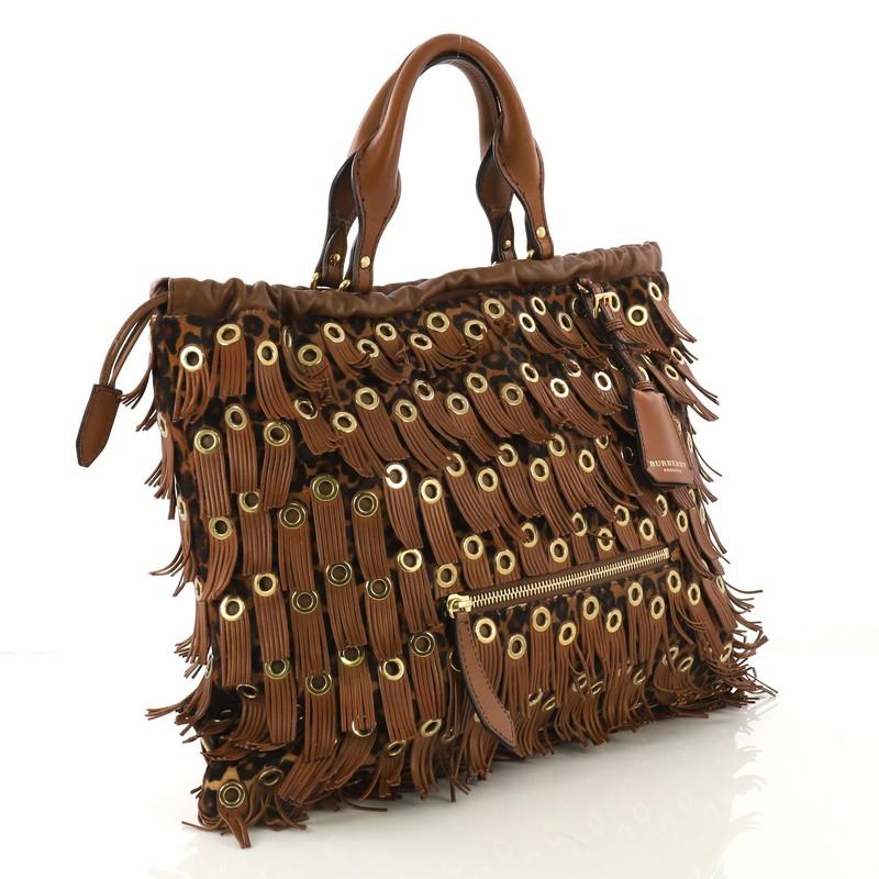 This Burberry Eyelets Big Crush Tote Fringe Calf Hair Large, crafted from brown calf hair, features dual rolled leather handles, fringe embellished with metal eyelets, exterior zip pocket, cinch closure at the top, and gold-tone hardware. Its