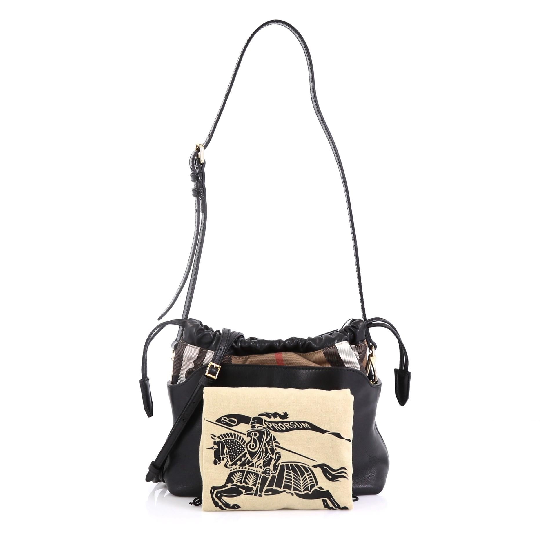 This Burberry Little Crush Crossbody Bag Leather and House Check Canvas, crafted from black leather and house check canvas, features an adjustable crossbody strap, polished Burberry logo plaque, and gold-tone hardware. Its drawstring and magnetic