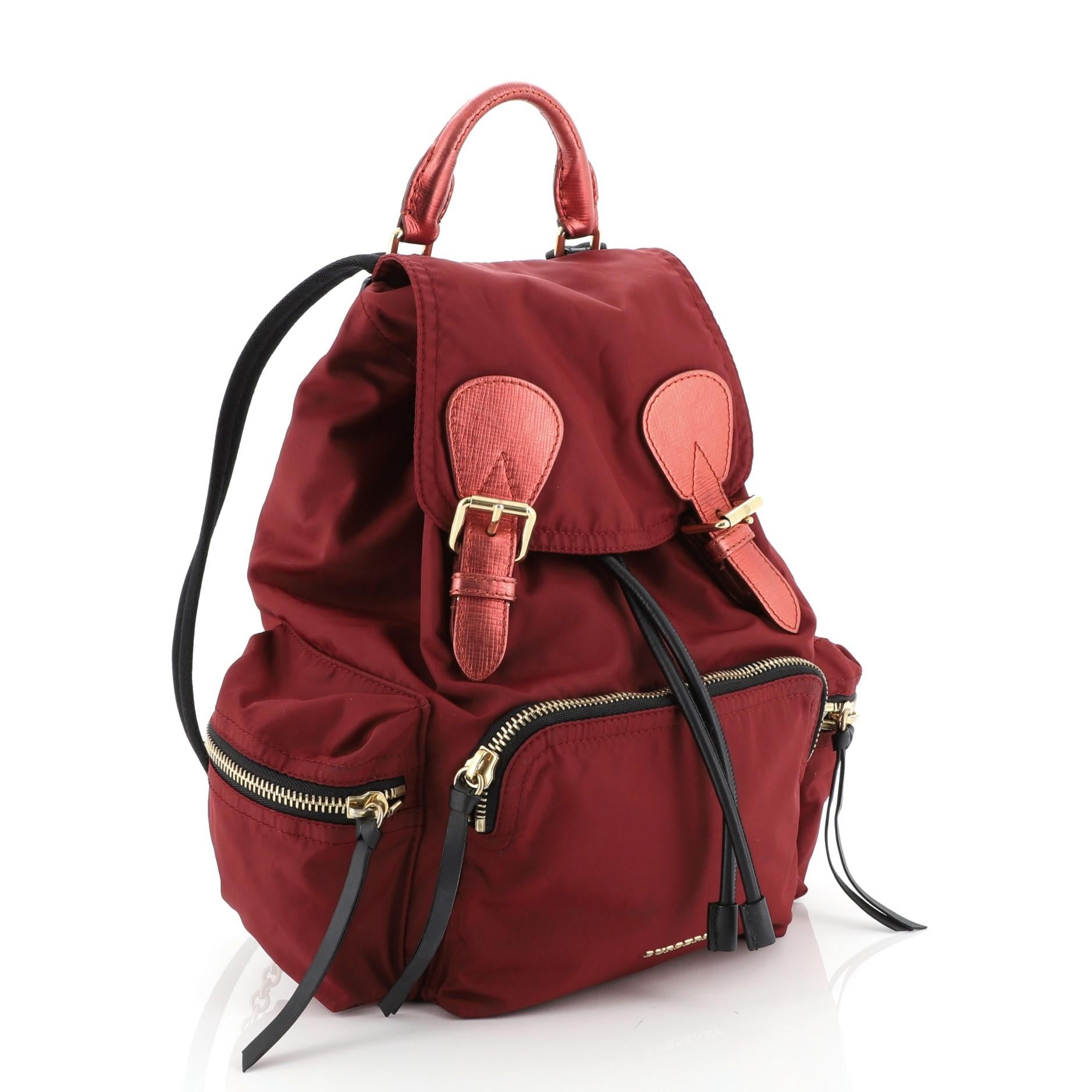 This Burberry Rucksack Backpack Nylon with Leather Medium, crafted from red nylon and leather, features a leather top handle, exterior zip pockets, quilted back panel, and gold-tone hardware. Its drawstring under snap-tab flap closure opens to a