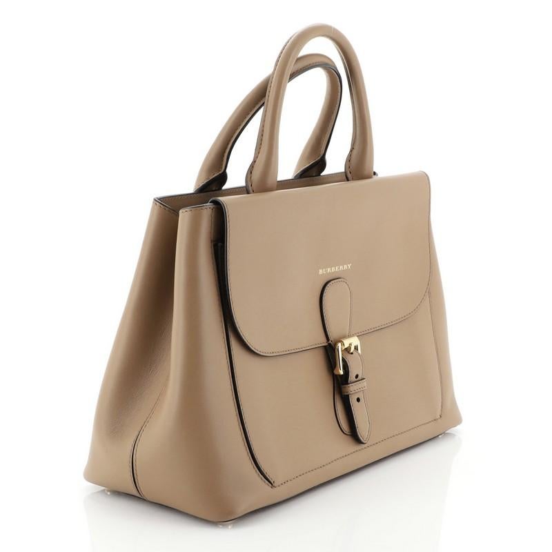 This Burberry Saddle Convertible Satchel Leather Medium, crafted in brown leather, features a sturdy, boxy silhouette, dual-rolled handles, front flap with belt buckle details, detachable strap, gold logo lettering, and gold-tone hardware. Its