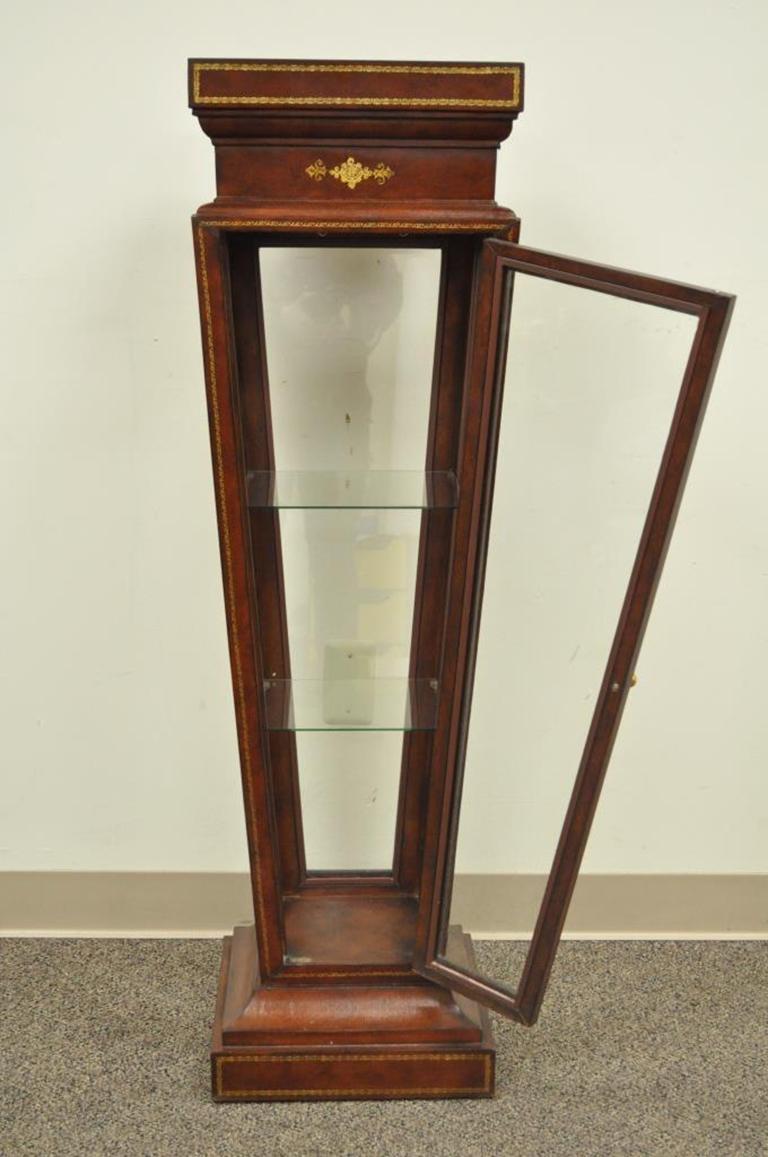 Unique leather wrapped pedestal or curio. Item features clear glass display case base with swing door, two interior glass shelves, beautiful tooled leather wrapped frame with gold gilt detailing, the maker is unconfirmed but possibly Maitland-Smith,