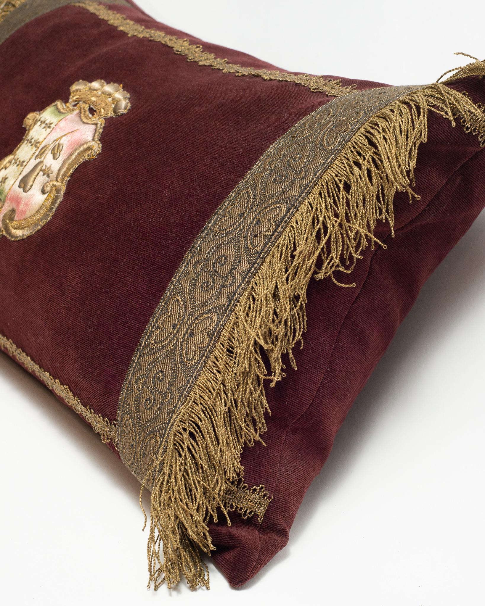 A burgundy velvet pillow with antique metallic gimp and large metallic crest.