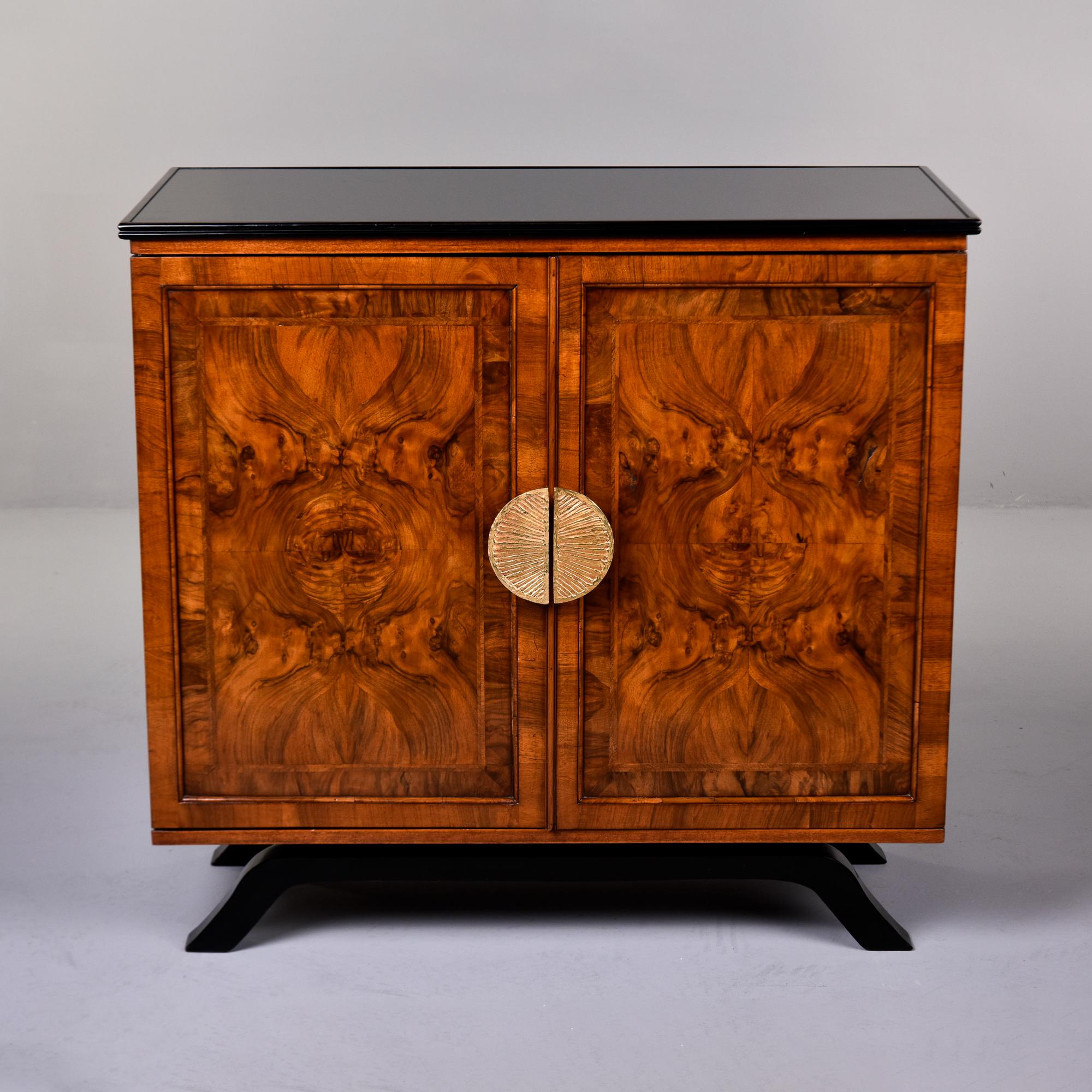 Found in France, this burl elm two door cabinet dates from the 1930s. Black glass top, burl elm veneer on the front and sides, original decorative brass handles and black footed base. Interior has adjustable shelves. Unknown maker. 

Very good