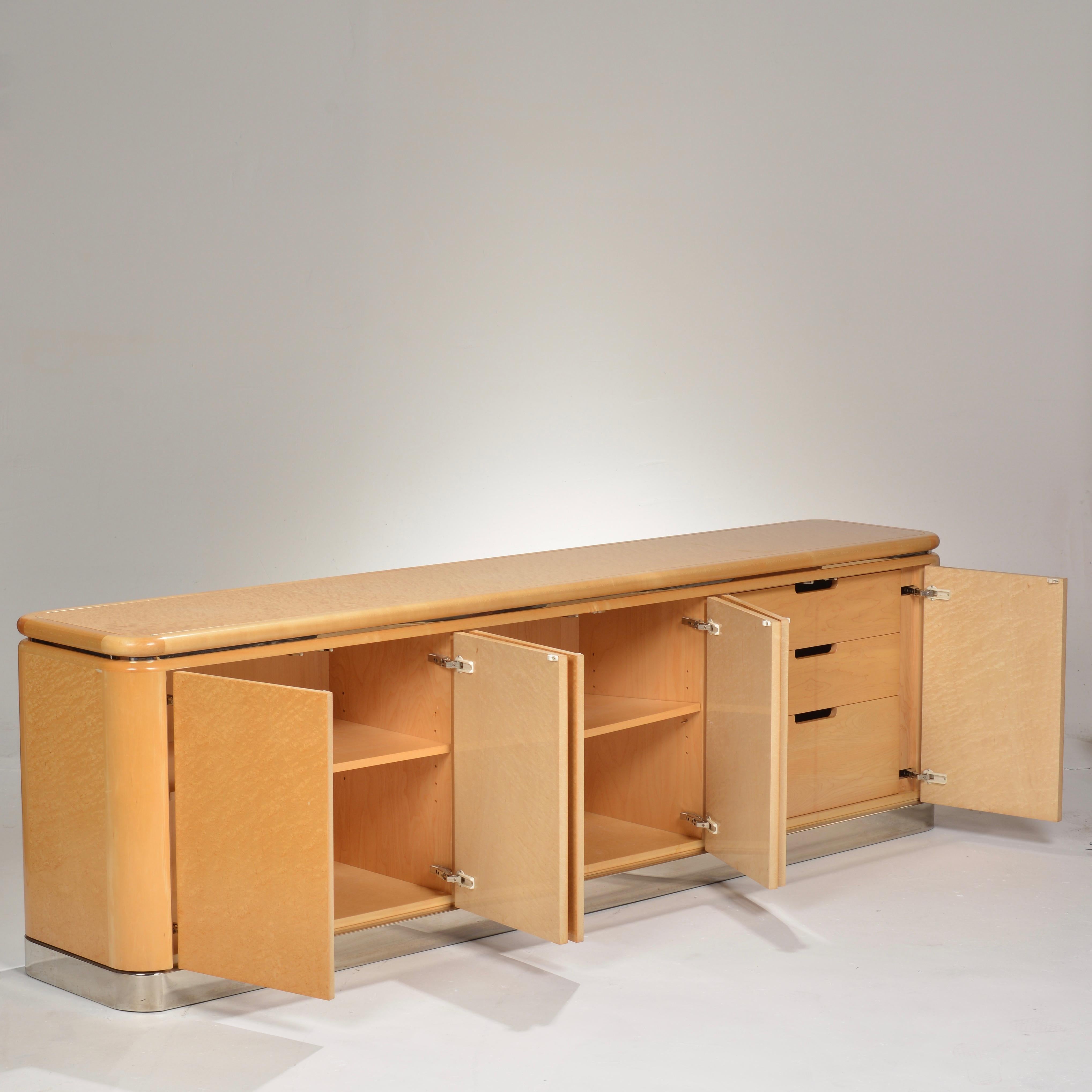 A beautiful 8' long credenza designed by Stanley Jay Friedman for Brueton featuring beautifully selected fiddleback maple handmade cabinetry, a polished stainless steel base, six doors, adjustable shelves, storage drawers and a finished back. This
