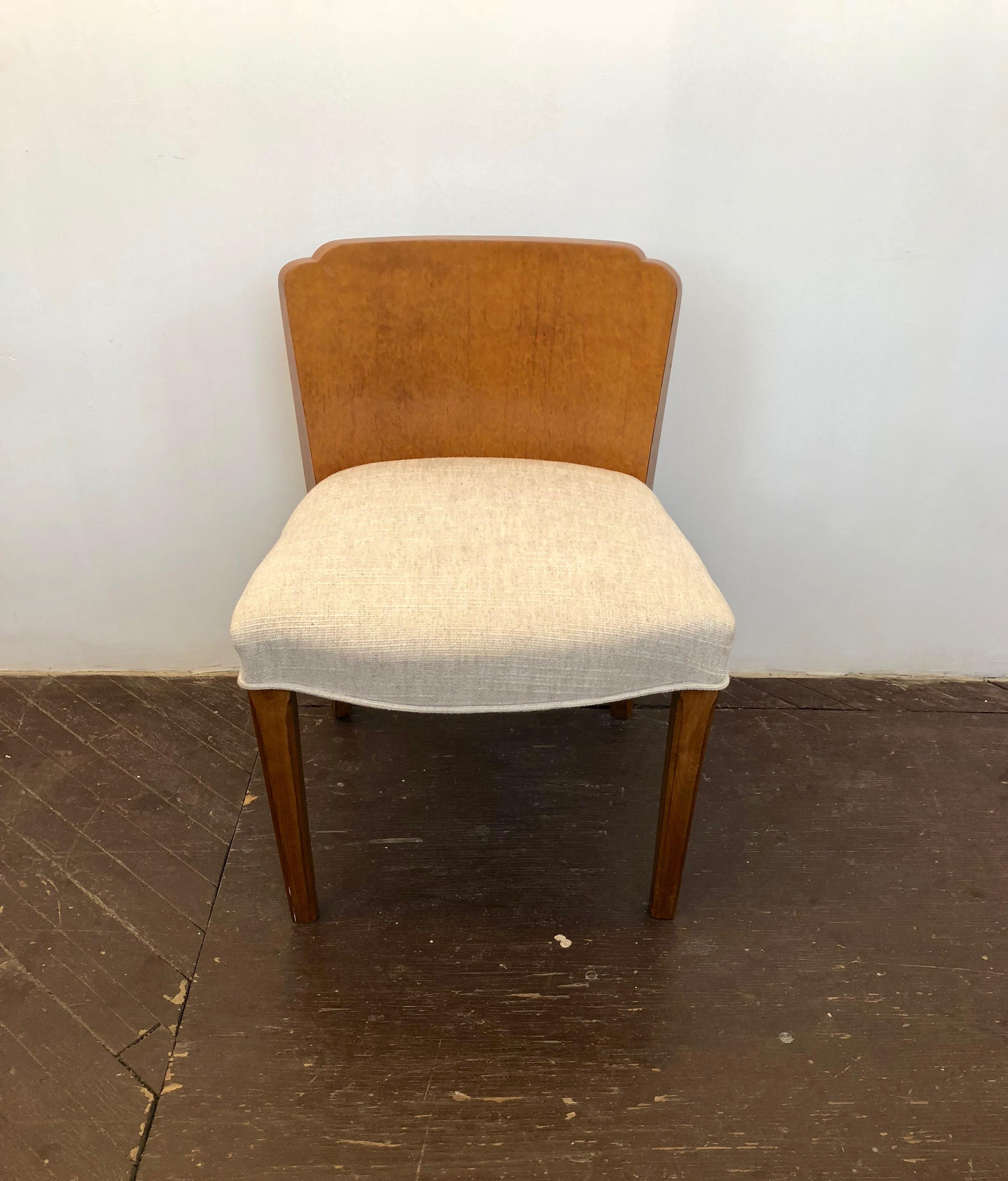 Restored American deco vanity chair has a burl wood back that has scalloped corners along the top, two wooden, arched, back legs and two wooden front legs that have a vertical carved detail along the edges, and a reupholstered curving seat in an