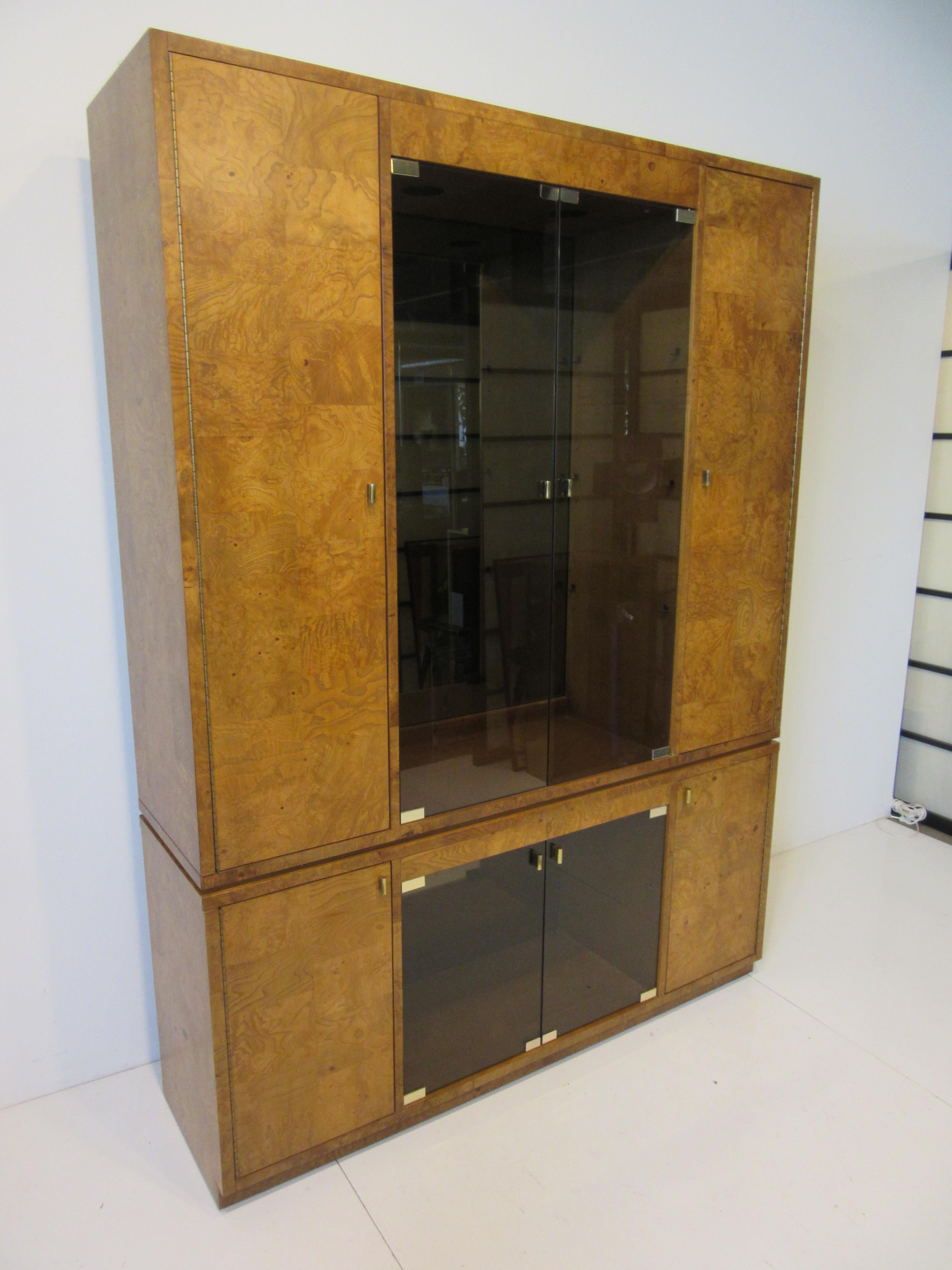 A very well constructed two piece wall unit / cabinet or bookcase in a medium toned burl wood having smoked glass doors and brass pulls and brass piano hinges. The upper piece has three adjustable glass shelves and to each side doors with three