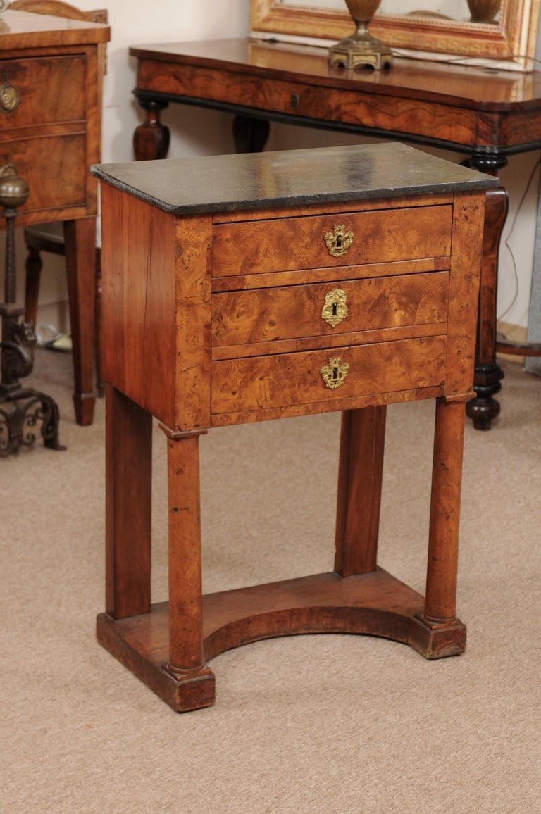 A burled elm Empire peite commode with black rectangular top, 3 drawers below with bronze escutcheons. All supported by turned column front legs and rectangular back legs ending in convex shaped base.
 