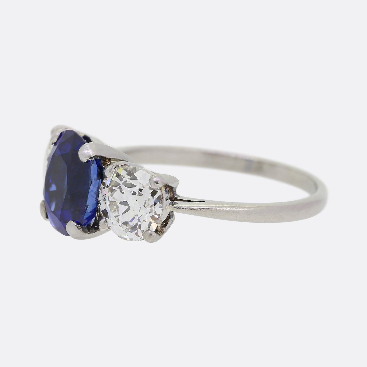 Nous avons ici une bague classique à trois pierres en saphir et diamant. Cette pièce ancienne met en valeur un saphir naturel de Birmanie au centre de la face, d'un bleu moyen intense. Cette pierre centrale de taille coussin est flanquée de part et