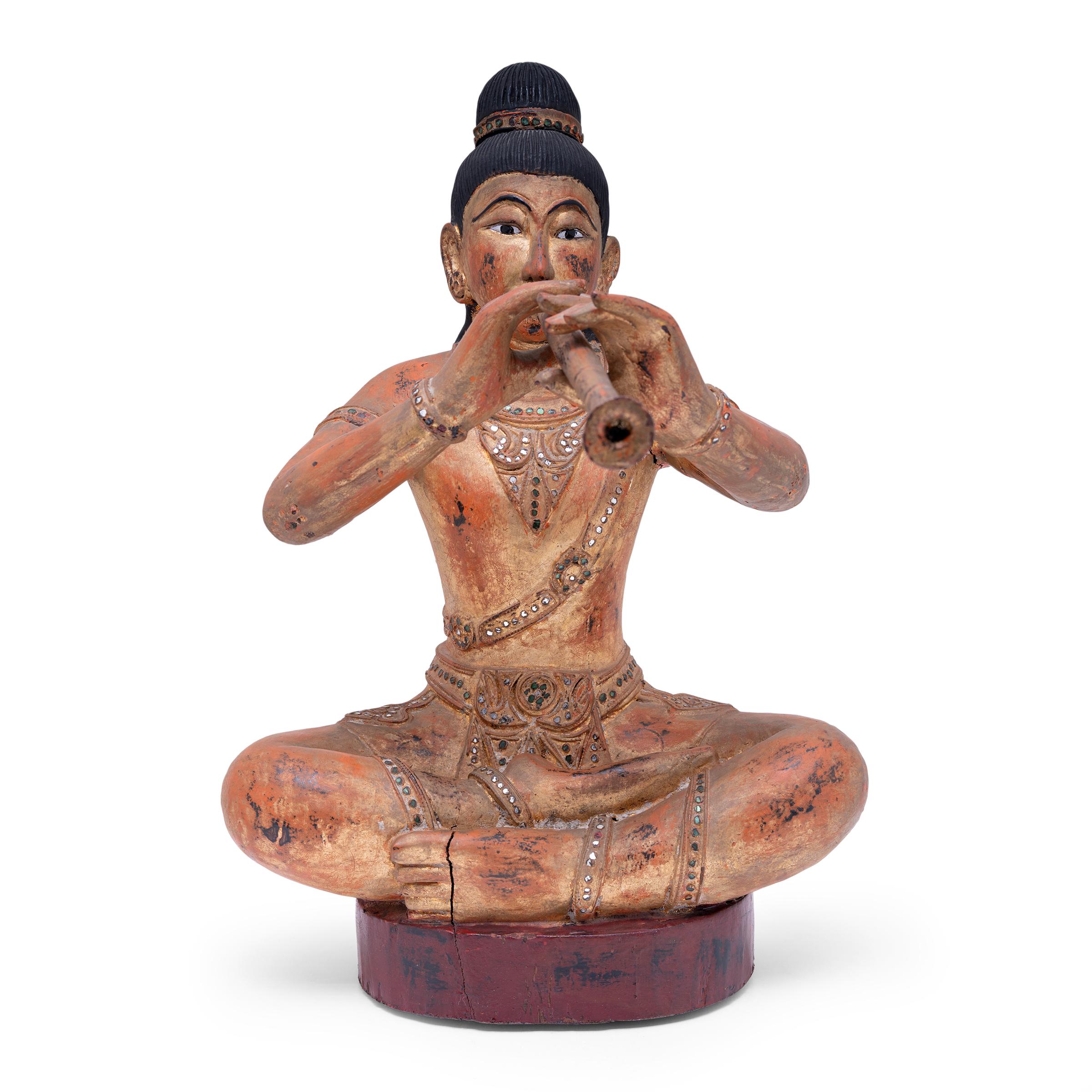 This seated Burmese figure captures a musician in eternal song. The wooden figure is hand-carved with a seated posture, legs crossed and arms outstretched to hold her instrument, most likely a flute or double-reeded horn. The musician is depicted in