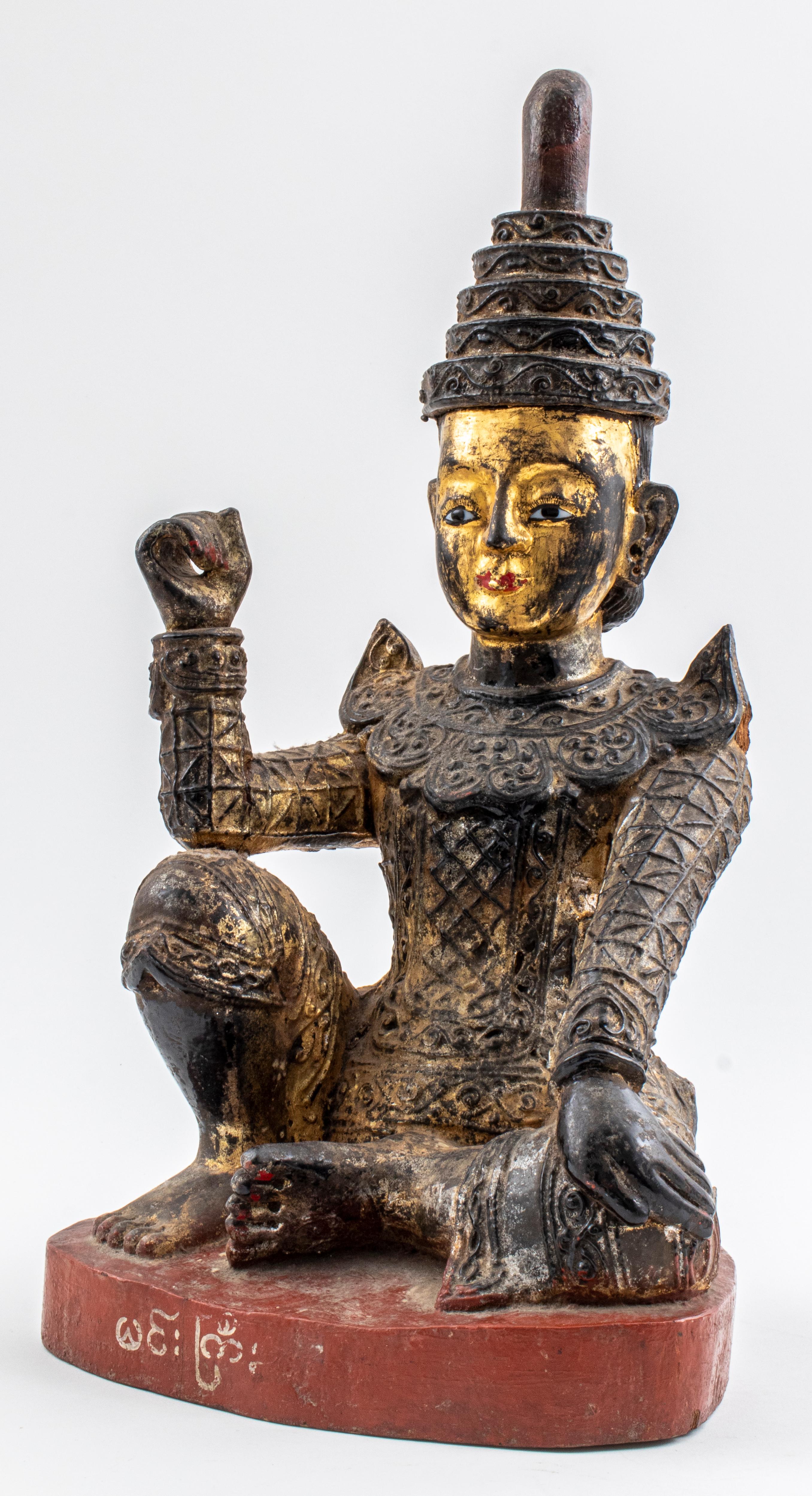 Statue ancienne en bois doré originaire de Birmanie représentant une figure de l'esprit NAT assise sur une base peinte en rouge avec des écritures effacées, le genou et le coude droits pliés et la main fermée pour former un cercle, ayant peut-être