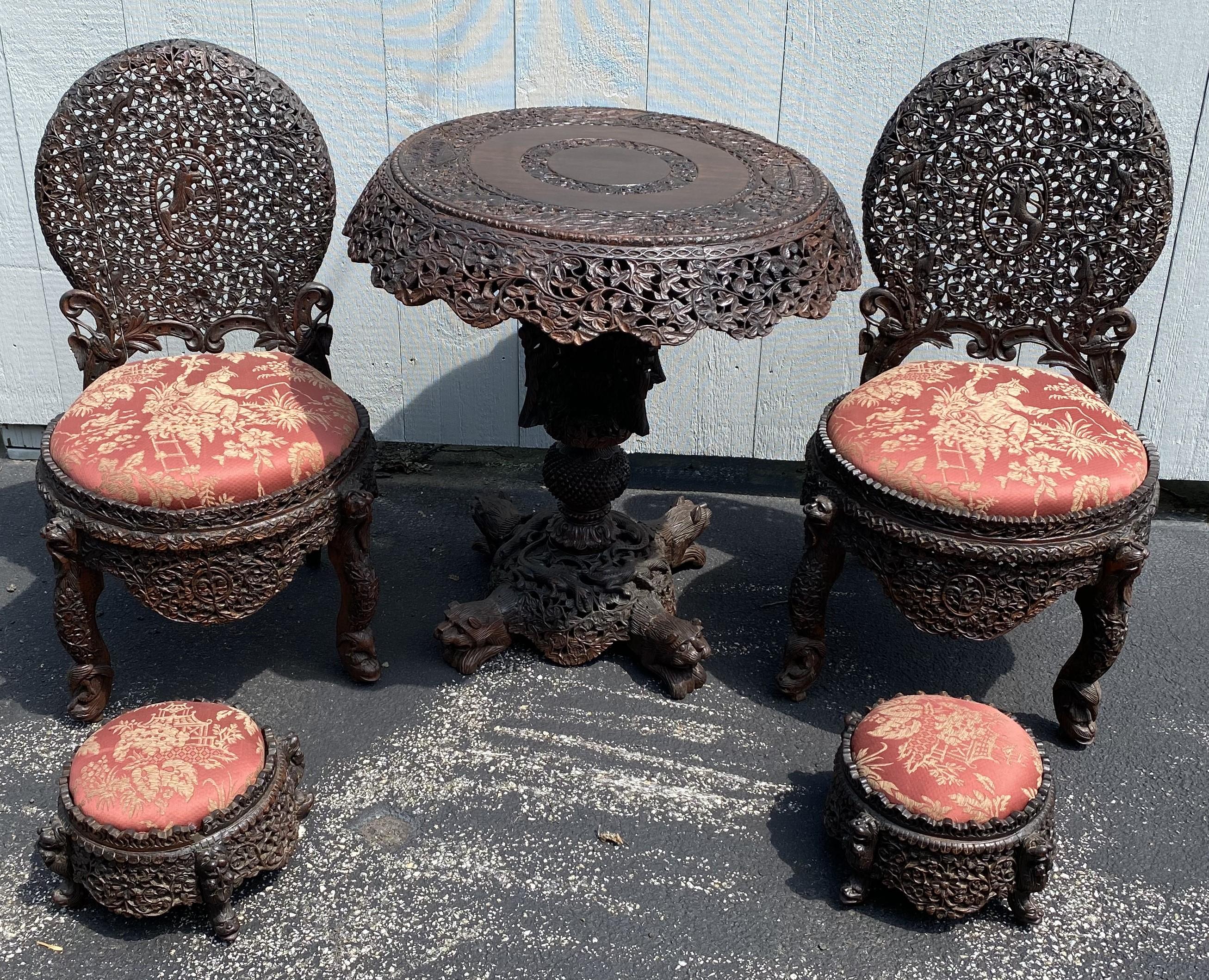 Magnifique ensemble en bois dur birman sculpté à la main, comprenant une table ronde à plateau basculant, deux chaises d'appoint assorties avec des jupes fortement sculptées et les poufs qui les accompagnent. Chaque pièce est sculptée à