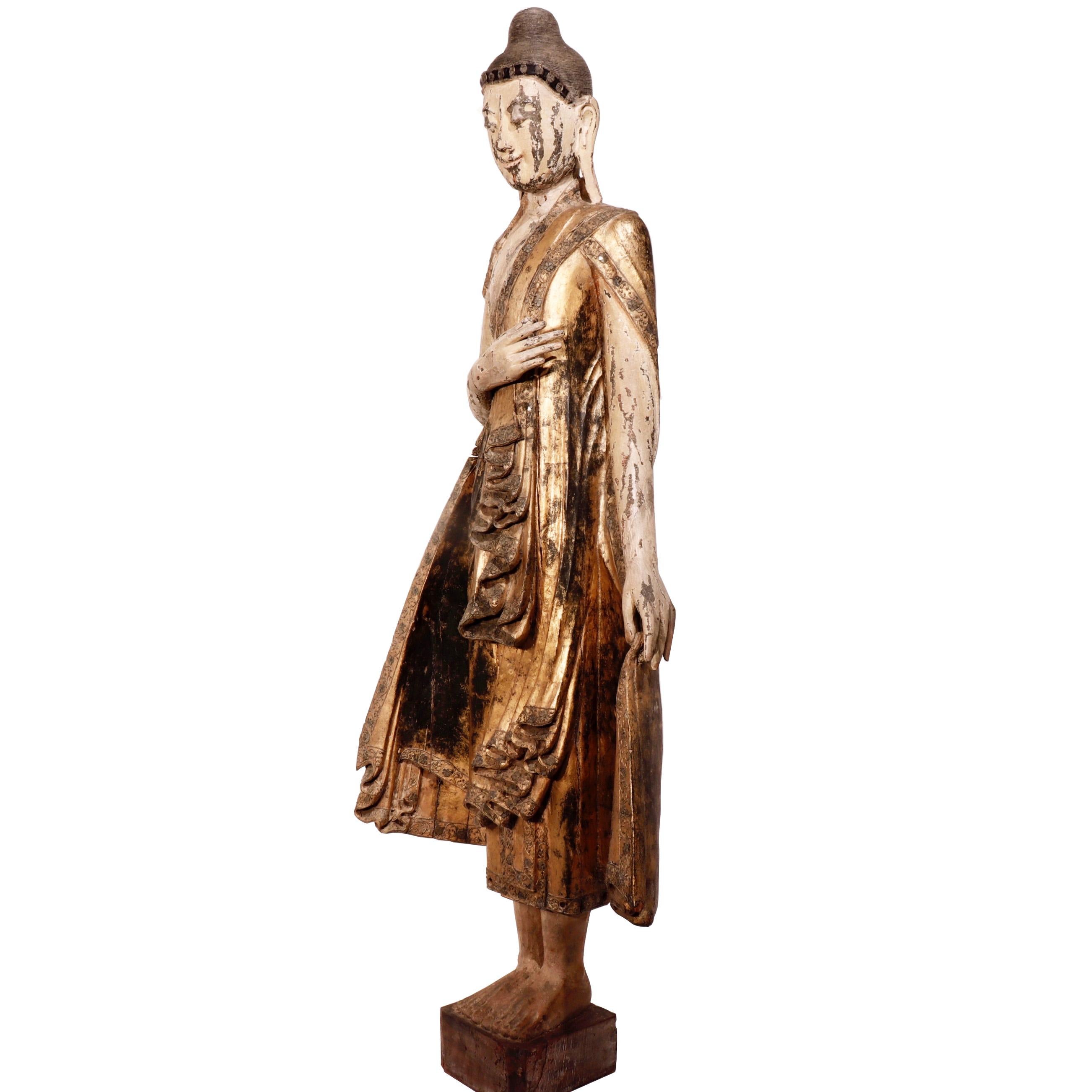 Figure birmane en bois sculpté de Mandalay représentant le Bouddha debout sur une base carrée, la main gauche dans le varada ou mudra d'octroi de bonté, la droite levée vers la poitrine, vêtu d'une robe décorée de bordures avec des inserts en verre