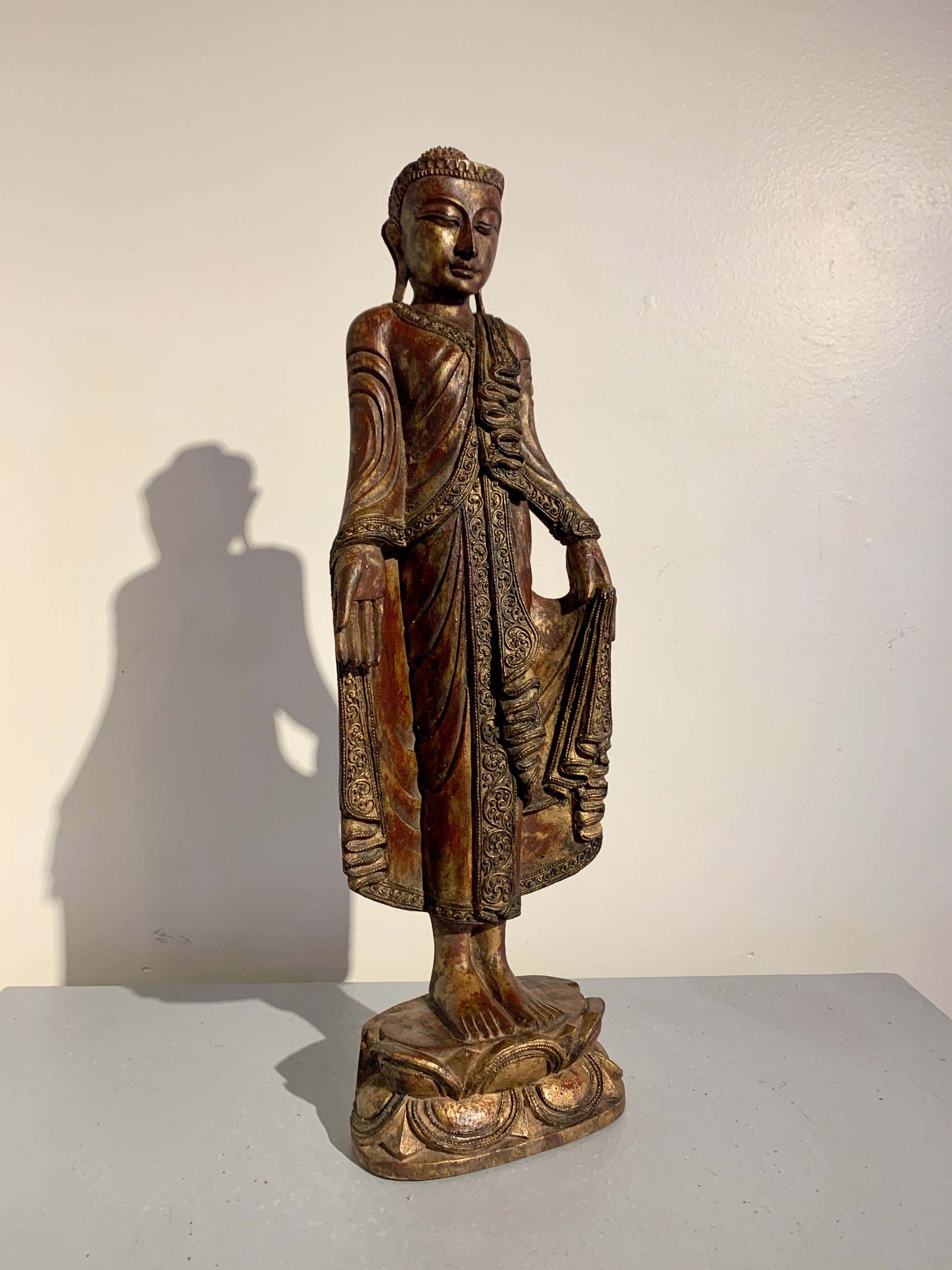 An enchanting Burmese carved hardwood, lacquered and gilt standing Buddha in the Mandalay style, early to mid 20th century, Myanmar (Burma). 

The regal Buddha is portrayed standing upon a stylized double lotus pedestal. He is dressed in heavy,