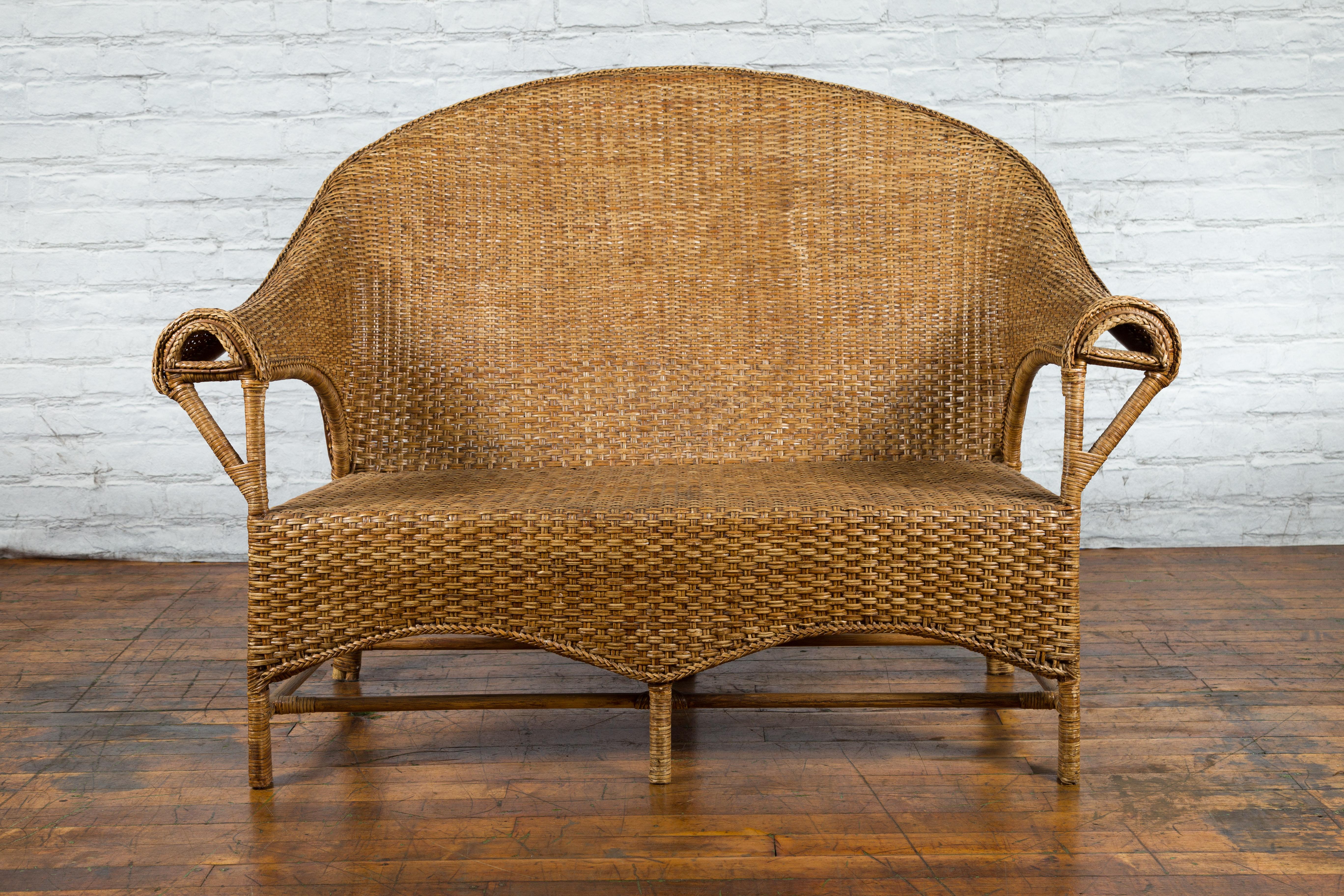 A vintage Burmese woven rattan and wood loveseat from the mid 20th century, with curving back, shaped skirt and side stretchers. Created in Burma during the Midcentury period, this charming wood and woven rattan loveseat features a curving back