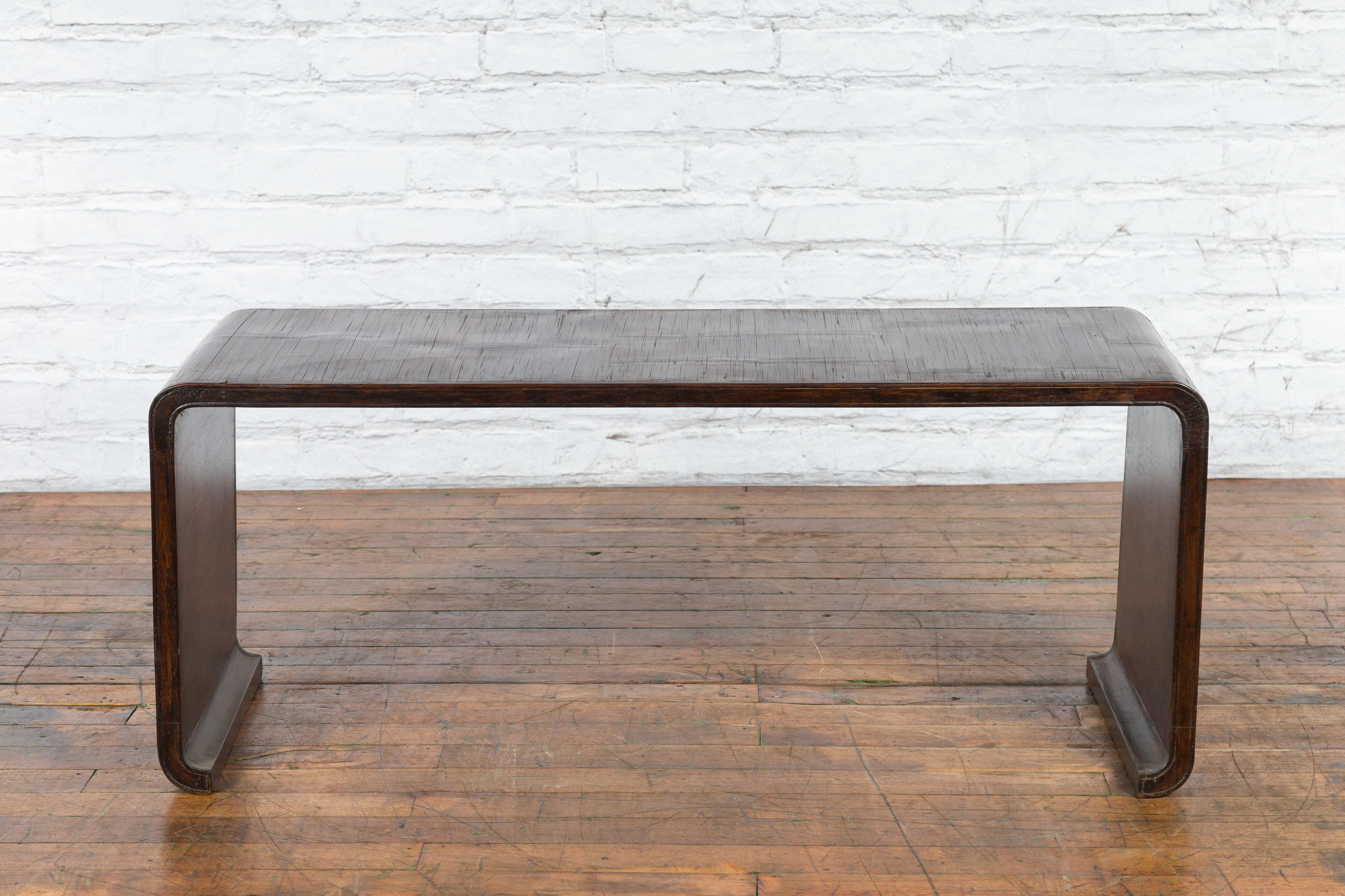 A vintage Burmese waterfall wooden coffee table from the mid-20th century with bamboo opium mat top and dark patina. Created in Burma during the midcentury period, this waterfall coffee table features a rectangular bamboo opium mat top flowing