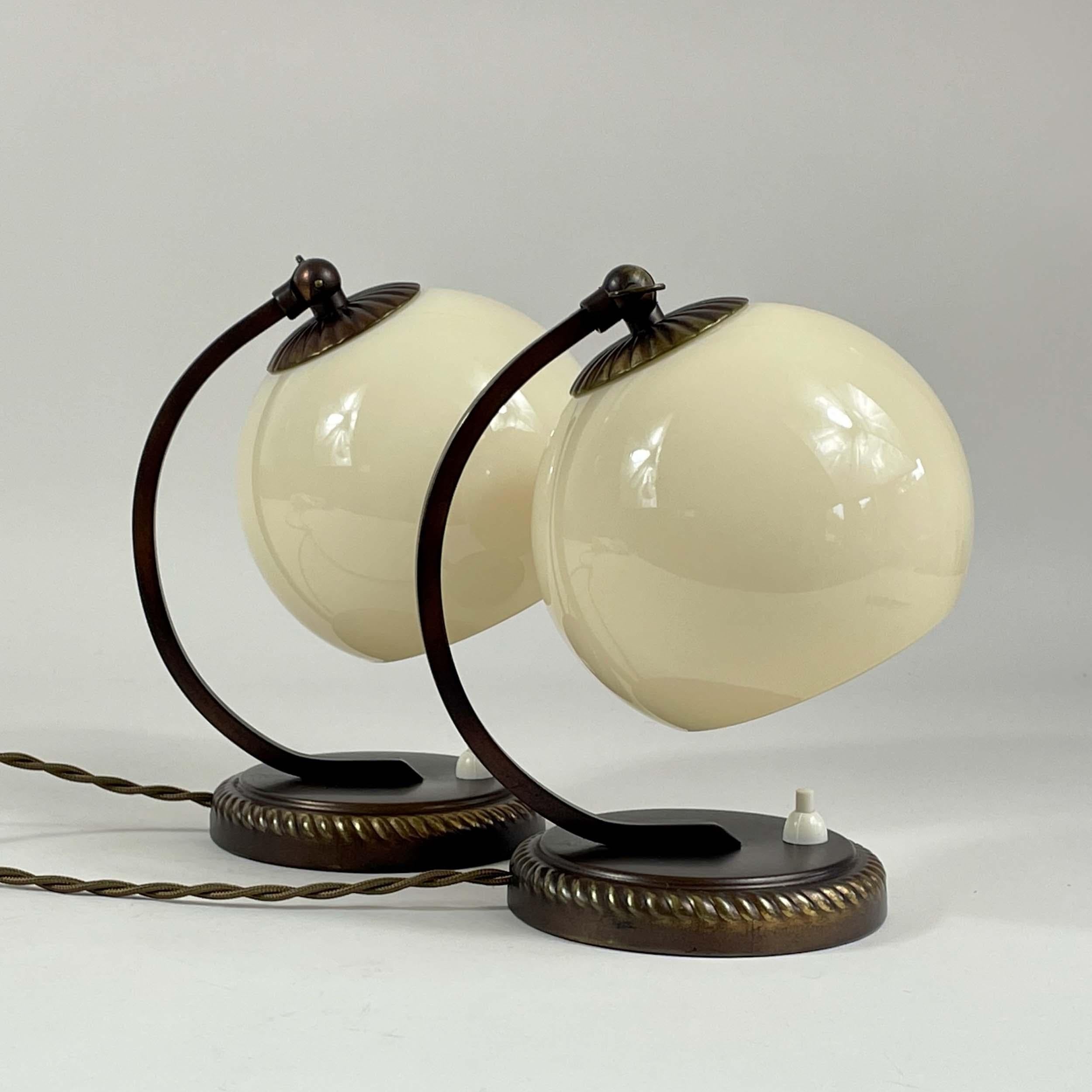 These table lights have been designed and manufactured in Germany in the 1930s to 1940s. 

The lights feature spherical cream-colored shades made of glossy opaline glass, supported by elegantly curved dark bronze arms that rise from circular bases