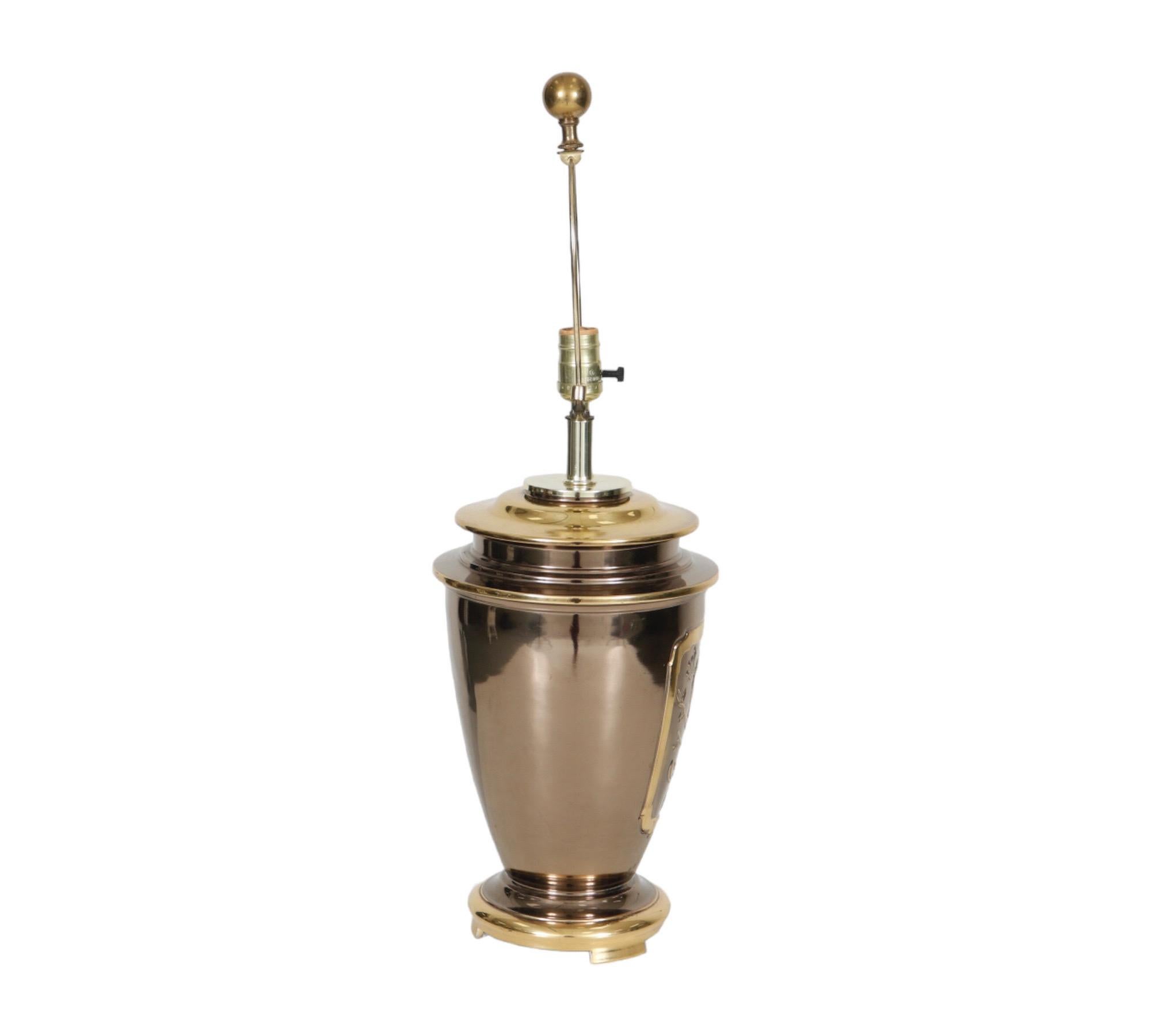 A burnished brass urn shaped table lamp. The front is decorated with an embossed shield that depicts a peacock on a branch mid-song. The round base is beveled with three block feet. Measure 9