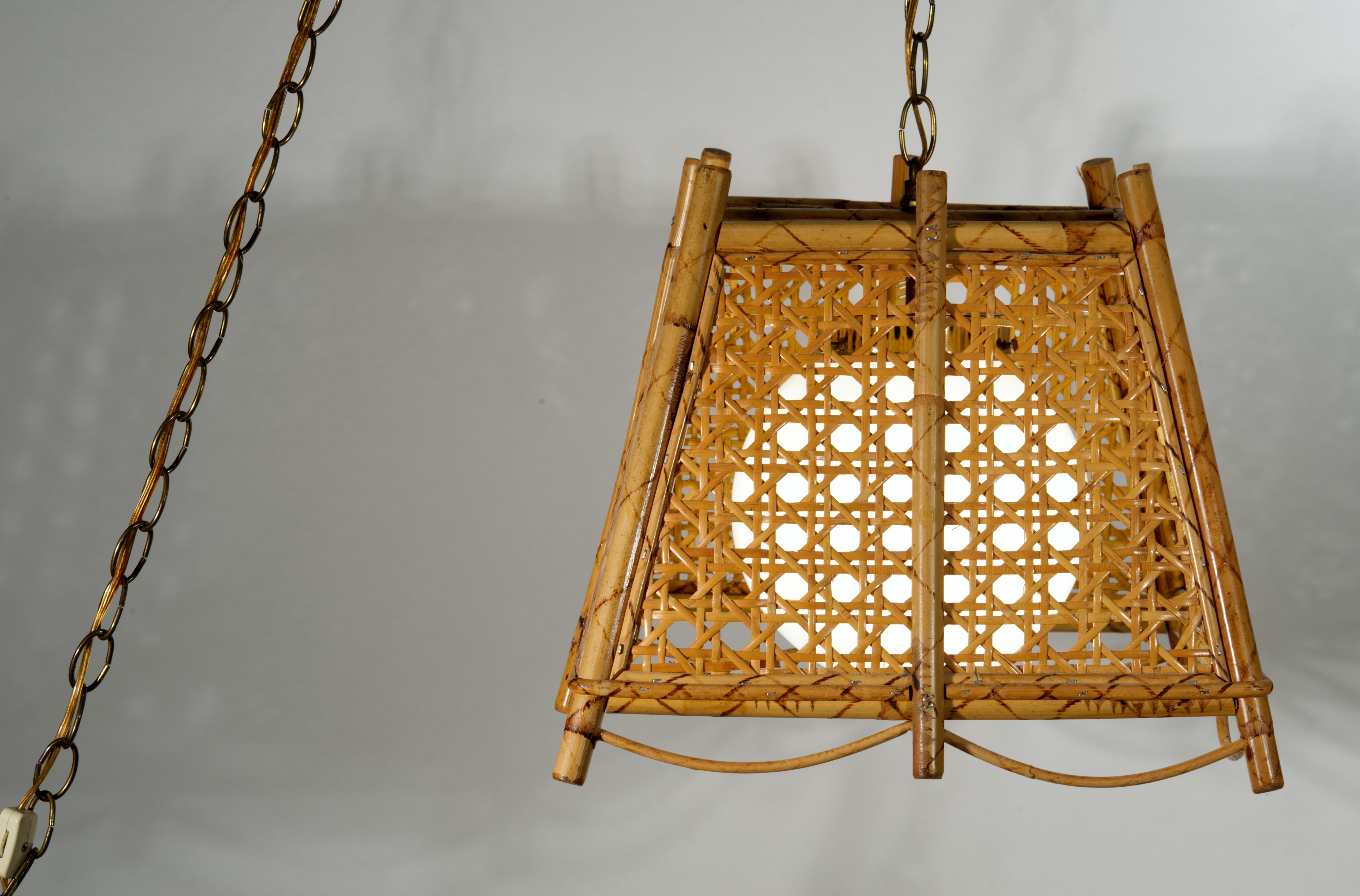 La lampada a sospensione o lampadario in bambù e vimini intrecciati della Modernità del Mid Century è caratterizzata da una struttura trapezoidale in bambù bruciato o tartarugato con intrecci geometrici in rattan che formano i lati del pendente; il