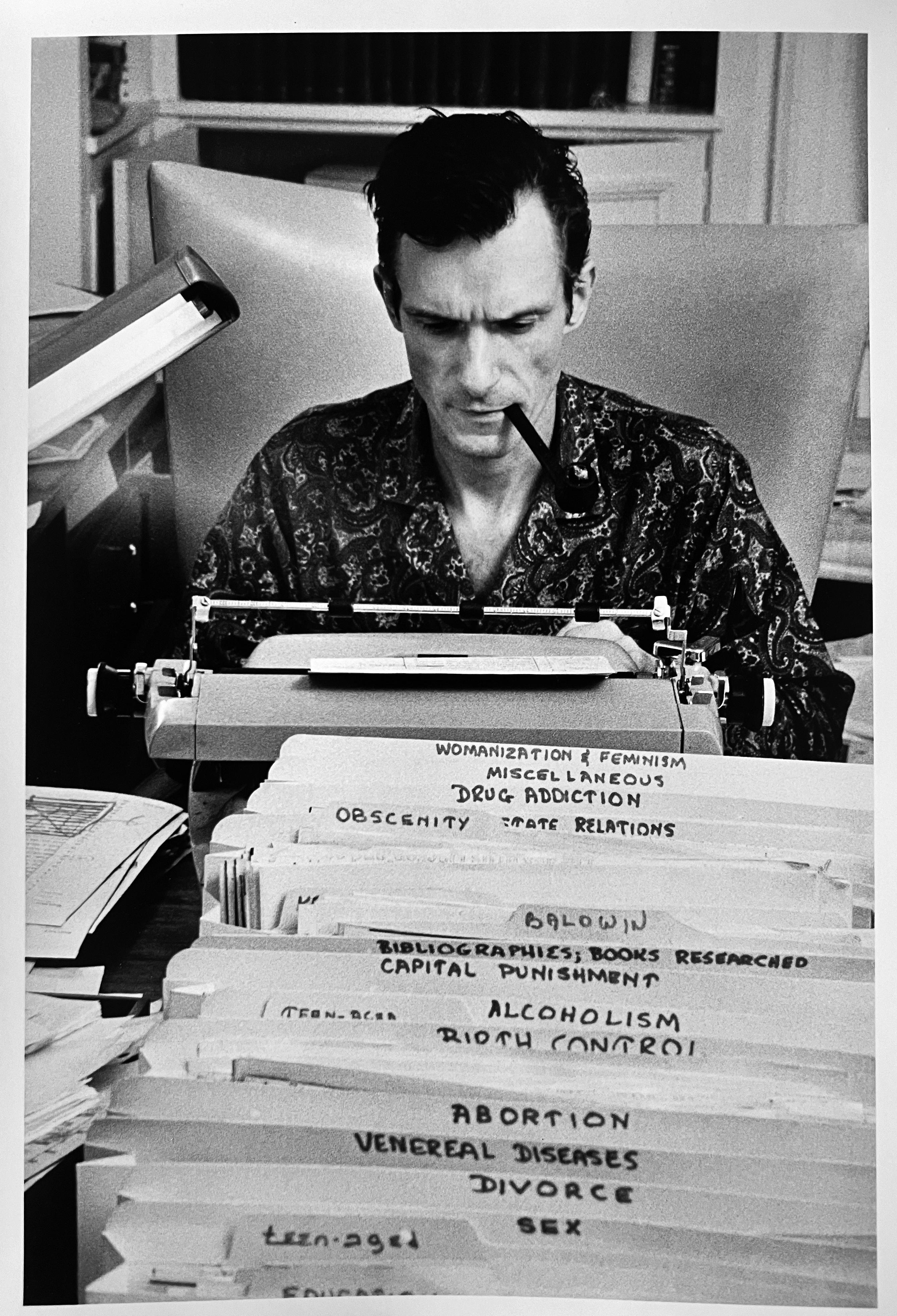 By American photographer Burt Glinn a black-and-white 14" x 11" photograph on fiber paper (gelatin silver print) of Editor-in-chief Hugh Hefner who wrote for the magazine he founded, Playboy. An historical photo of Hefner smoking a pipe at his desk