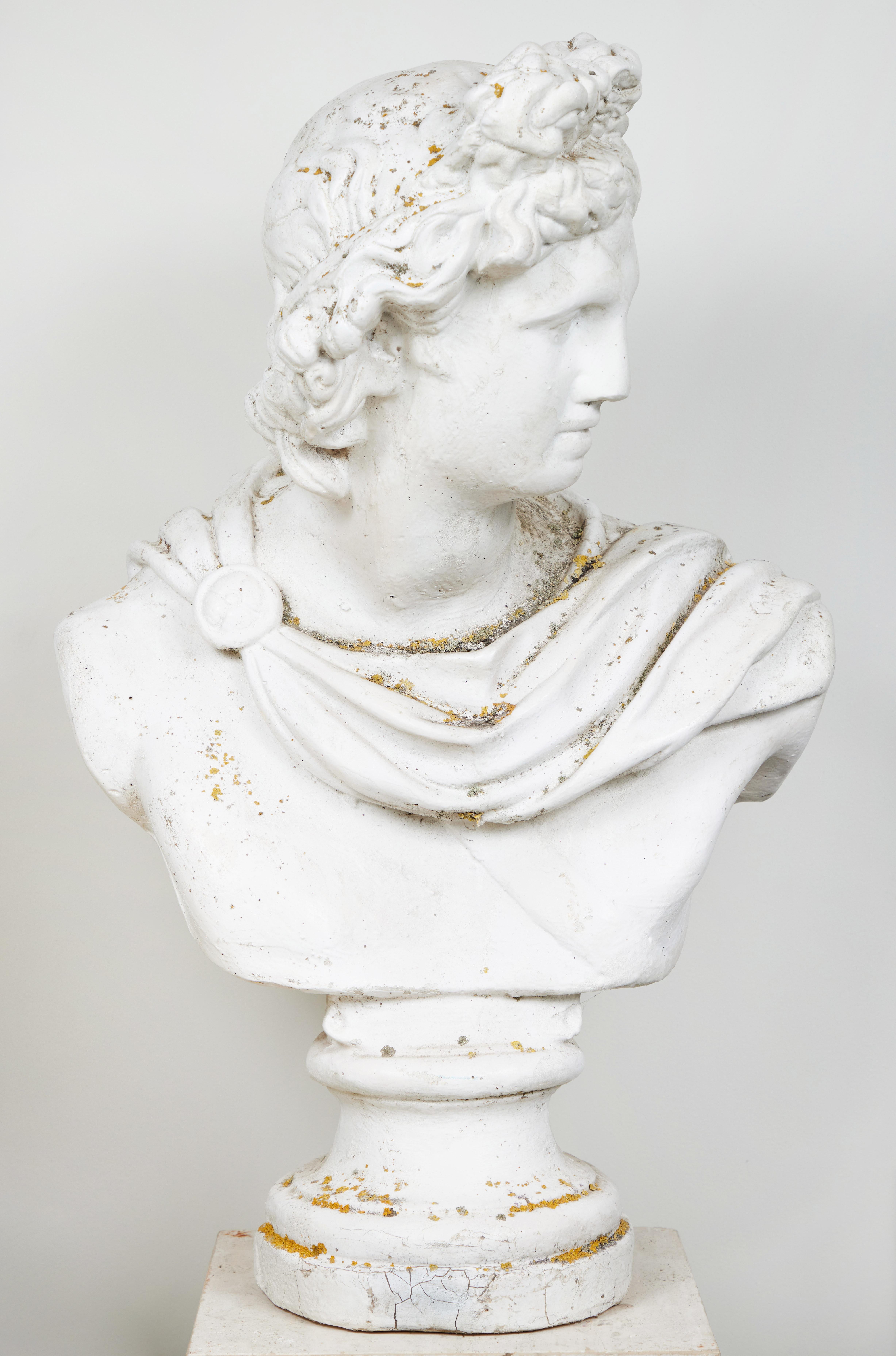 Cast stone garden statuary bust after the Apollo Belvedere, wearing a headpiece and draped gown. On a socle base.