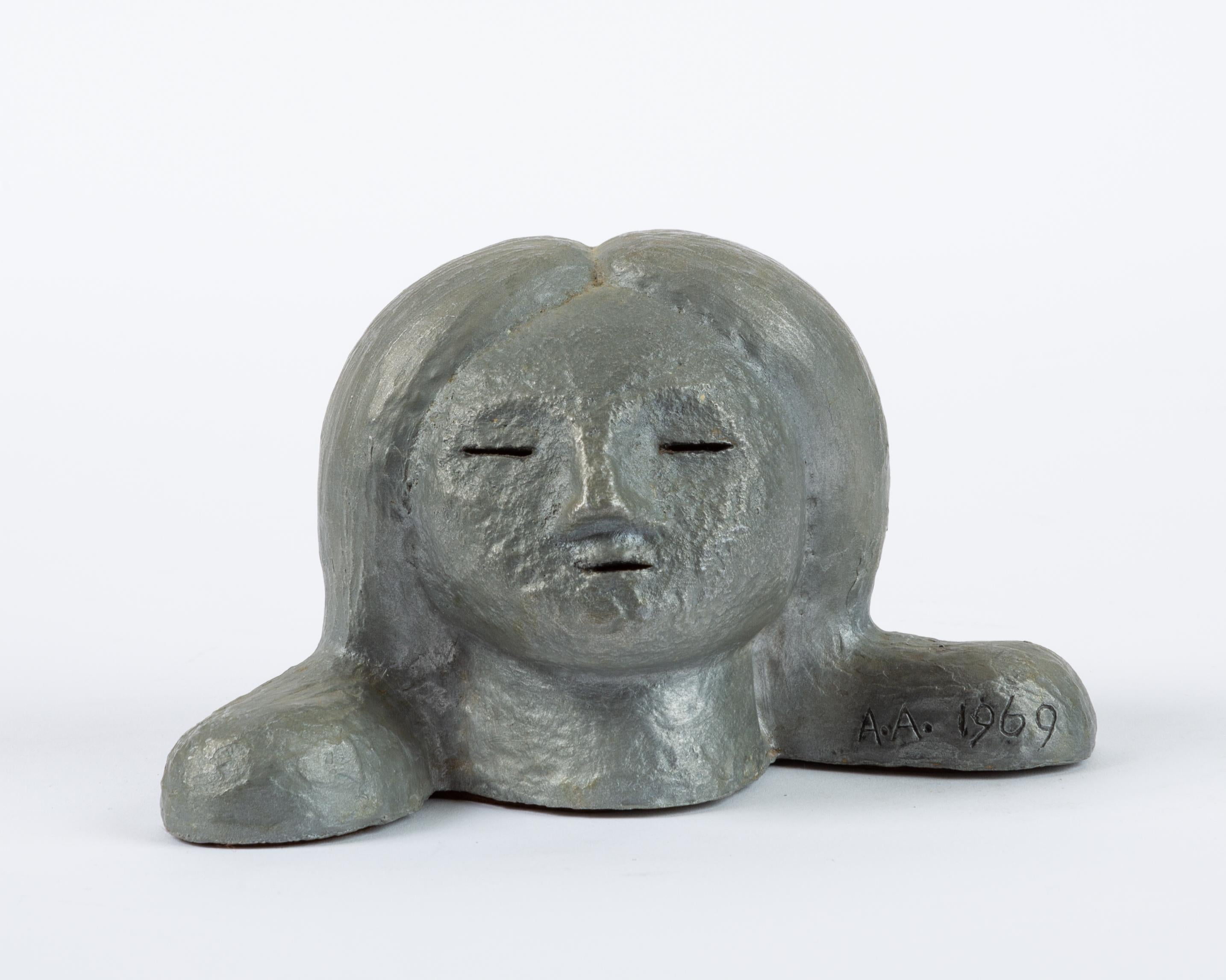 A small plaster head of a child with exaggerated pigtails, wide nose, and delicate eyes and mouth. Built over a papier-mâché structure, the figurine has been painted with a metallic silver enamel. Inscribed on foot, “Alexa Acuña August 16,