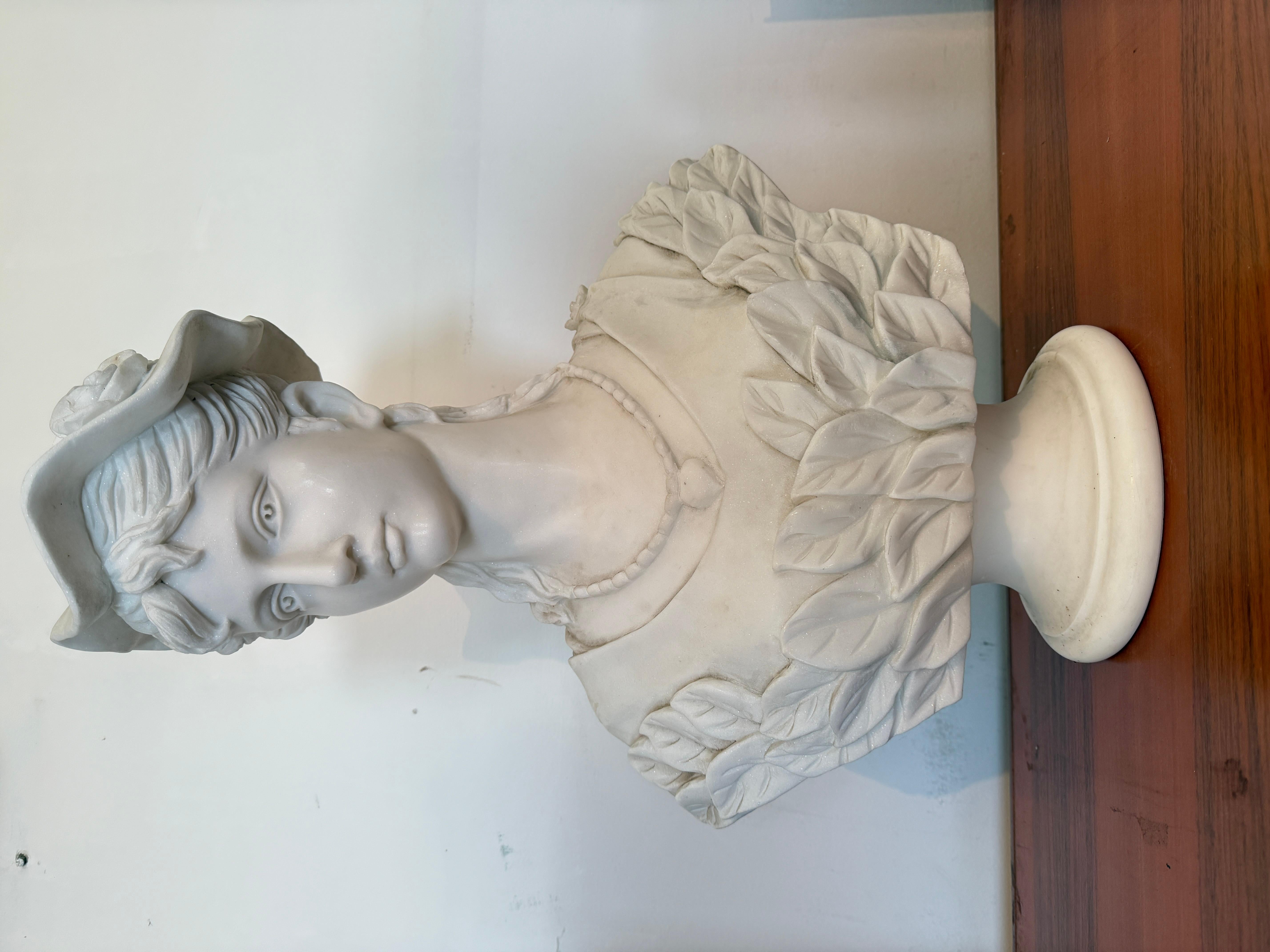 Raffinato busto in marmo bianco raffigurante una giovane donna con cappello ornato da fiori e abito decorato con un collier e un'elegante ghirlanda di foglie. L’opera cattura con grazia l’estetica dello stile Liberty, caratterizzata da linee