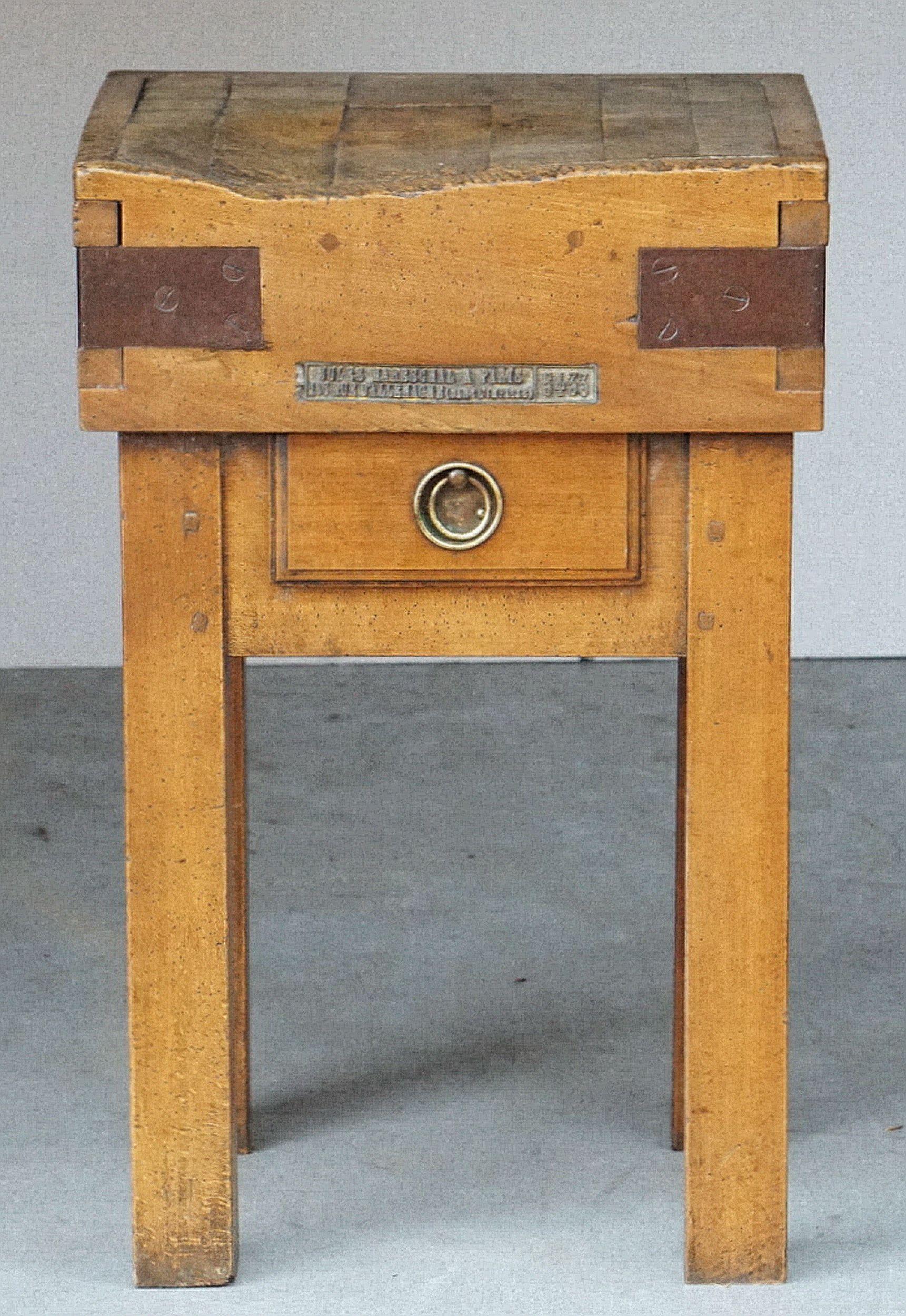A handsome French butcher's chopping block or table, from the 19th century, featuring a large, square sloping block or slab of iron-bound wood set upon a four-legged support stand of pine. Stand with drawer and ring pull.

With label to front: