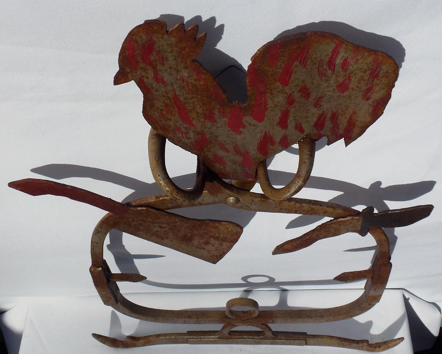 This is a folky butcher's trade sign. It is hand made from metal tools and found objects, with a sheet metal rooster on top. Ice tongs form part of the frame. It has worn silver paint with areas of surface rust. Red and black decoration was added on