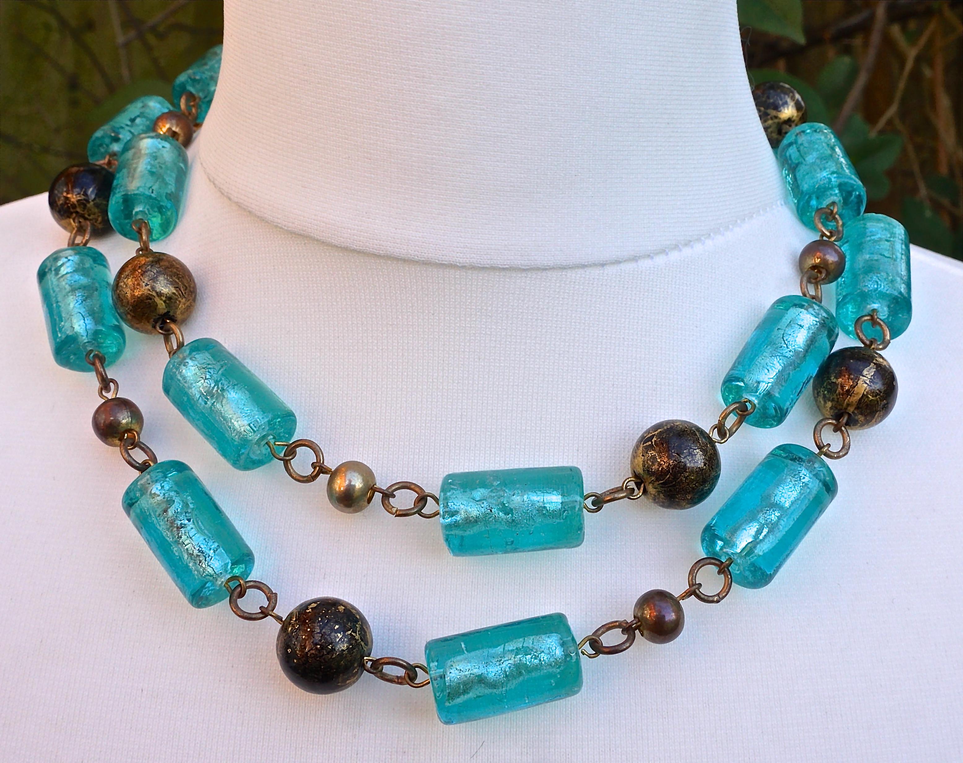 Beautiful Butler & Wilson gold tone double strand necklace, featuring cylinder shape aqua blue glass beads, black beads with a golden finish (possibly hand painted), and small metal beads. There is the Butler & Wilson tag on the end of a silver tone