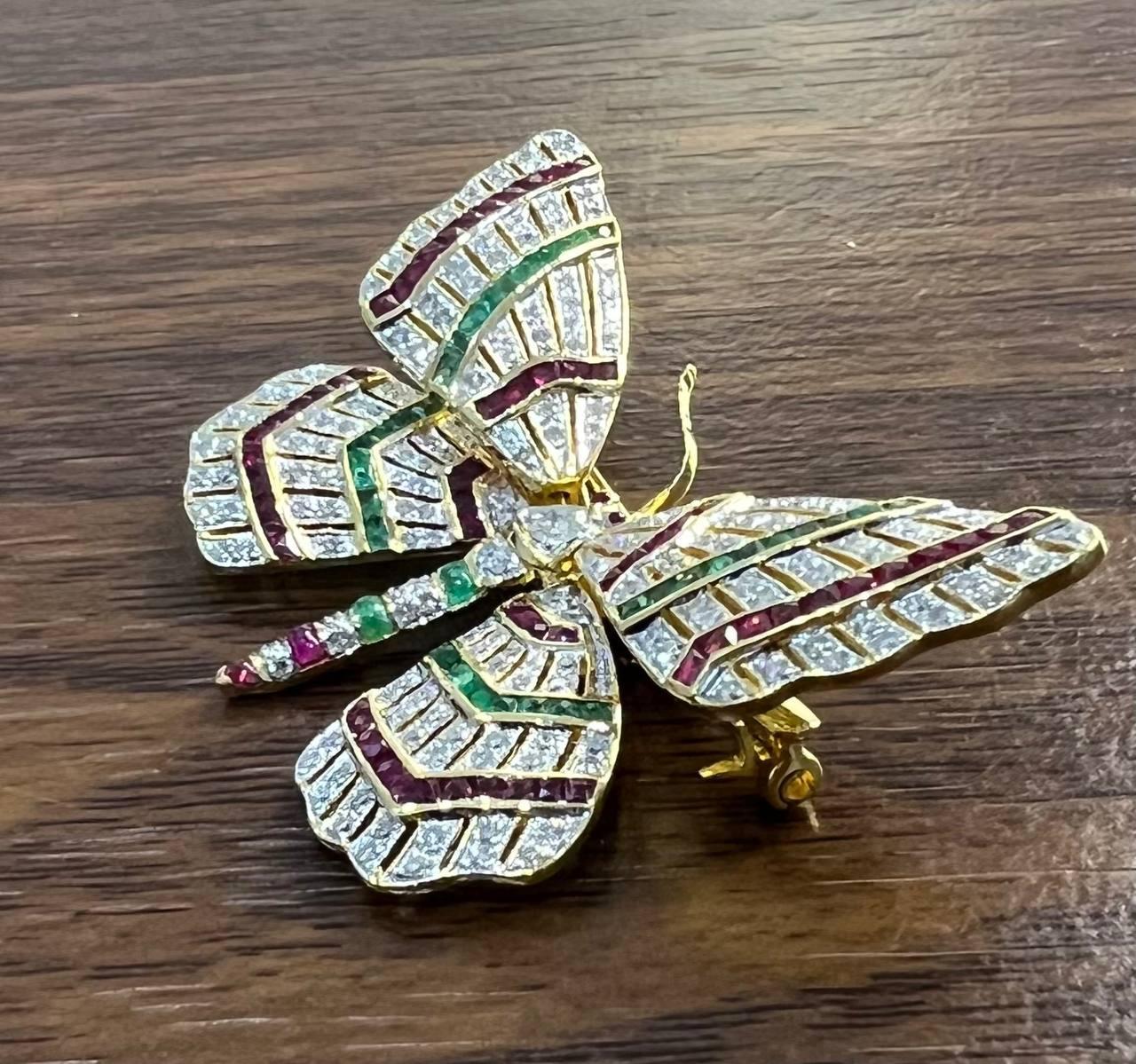 This exquisite brooch features a butterfly design adorned with vibrant rubies, emeralds and diamonds. The wings are intricately set with alternating rows of green, red, and white stones, creating a striking pattern. The body of the butterfly is