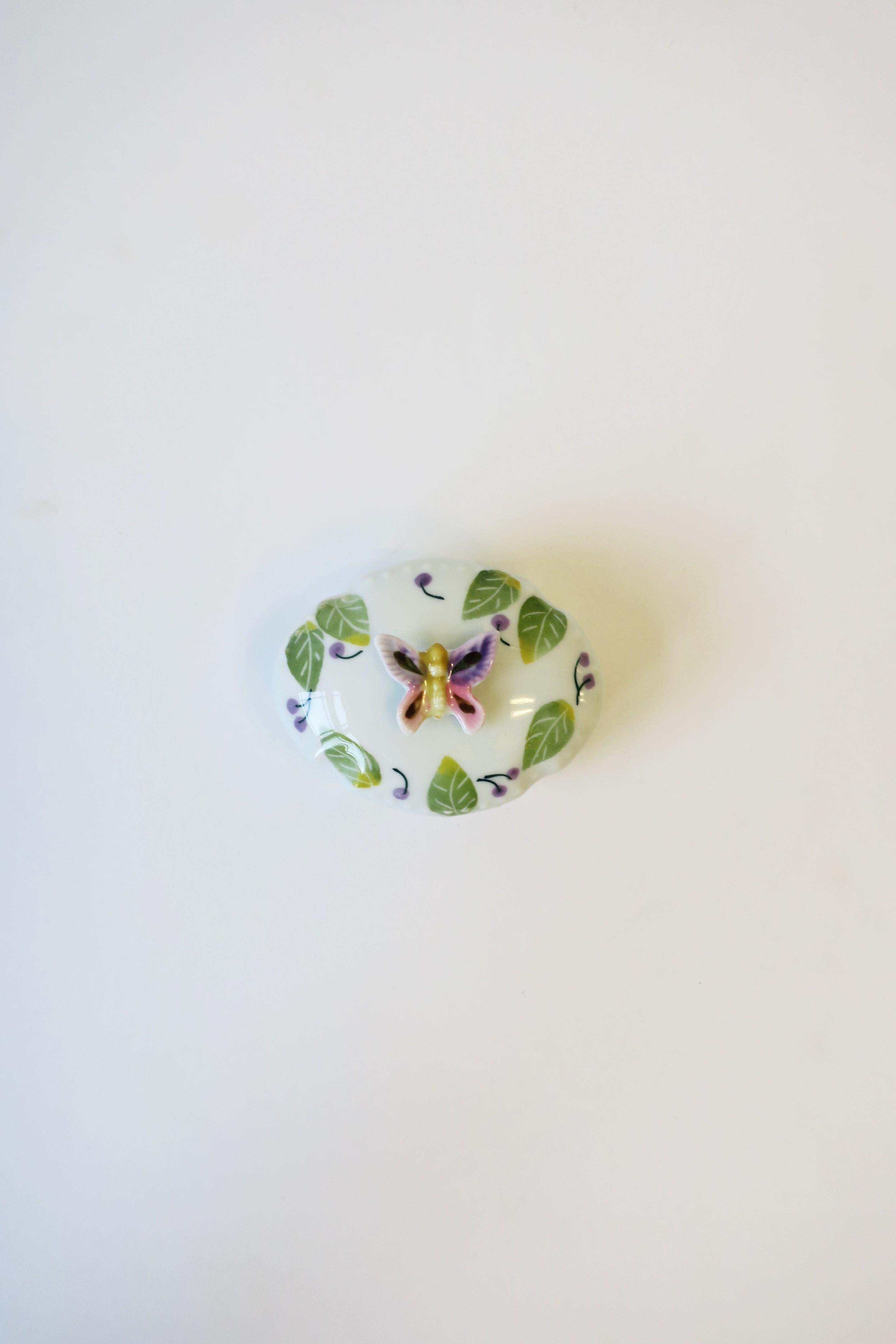 A beautiful small white porcelain oval jewelry or trinket box with colorful butterfly, leaves and cherries, circa mid to late-20th century. Piece has a raised butterfly knob on lid; an easy open close to place jewelry or other small items on a desk,