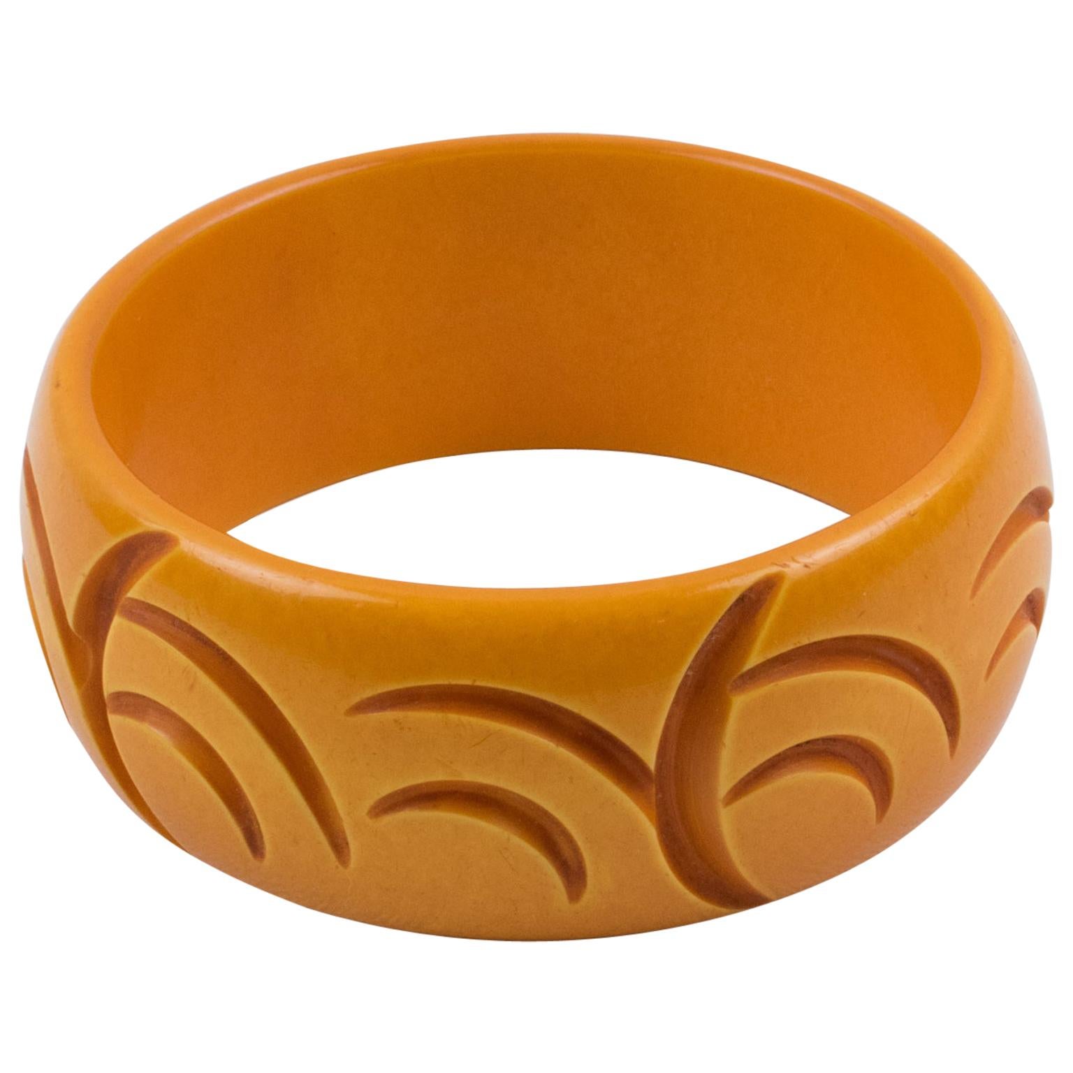 Questo affascinante bracciale bangle in bachelite color burro presenta una forma a cupola con profondi intagli geometrici tutt'intorno. Vanta un design a mezzaluna in un intenso colore giallo-arancio. 
Il bracciale è in buone condizioni, con leggeri