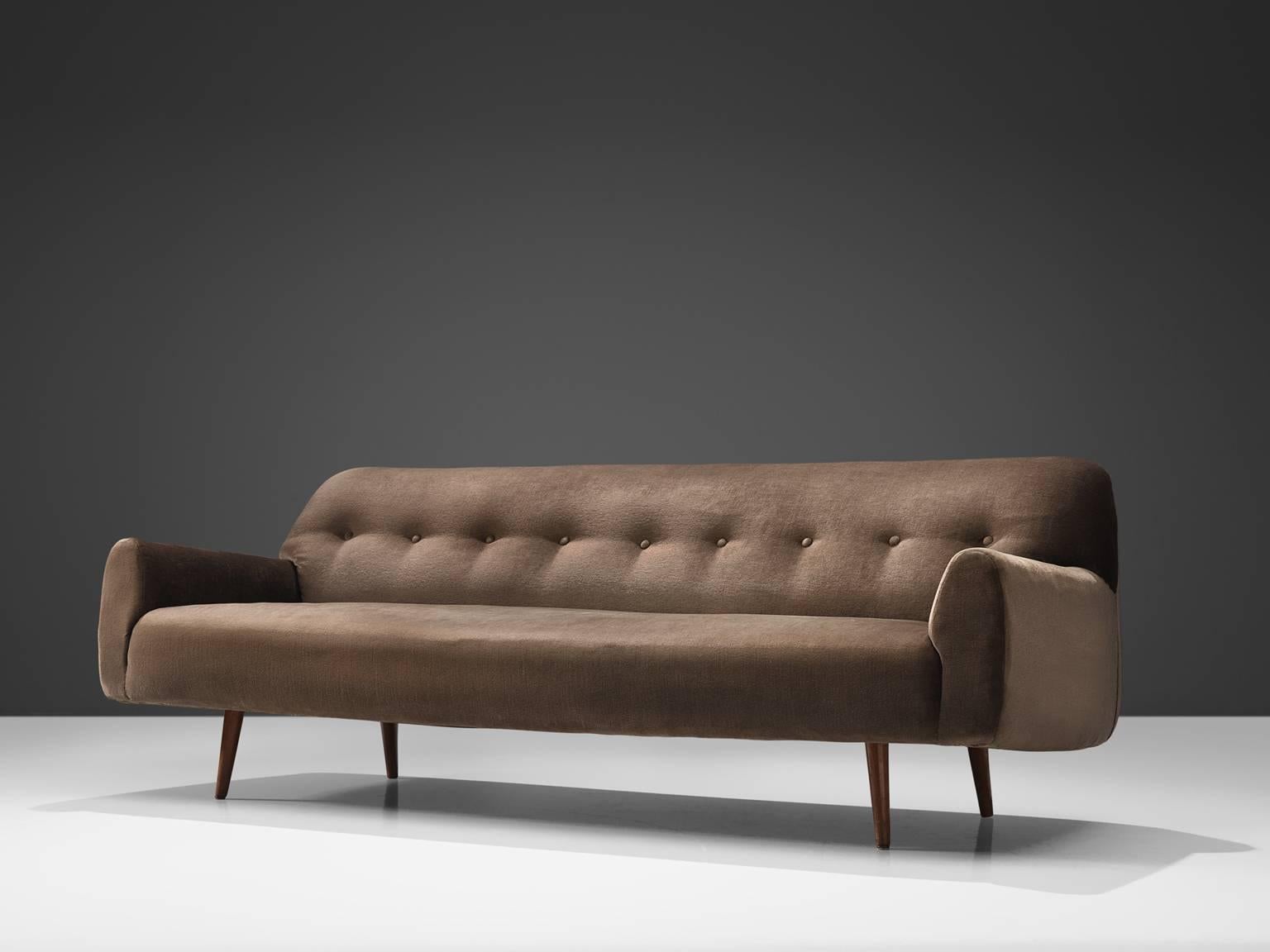 Sofa, velvet, wood, Europe, 1960s.

This large three-seat sofa features a dark wooden tapered conical, angled legs. The sofa is voluptuous and thick with rounded corners. The combination of the thin wooden legs and the thick bulky seat form a