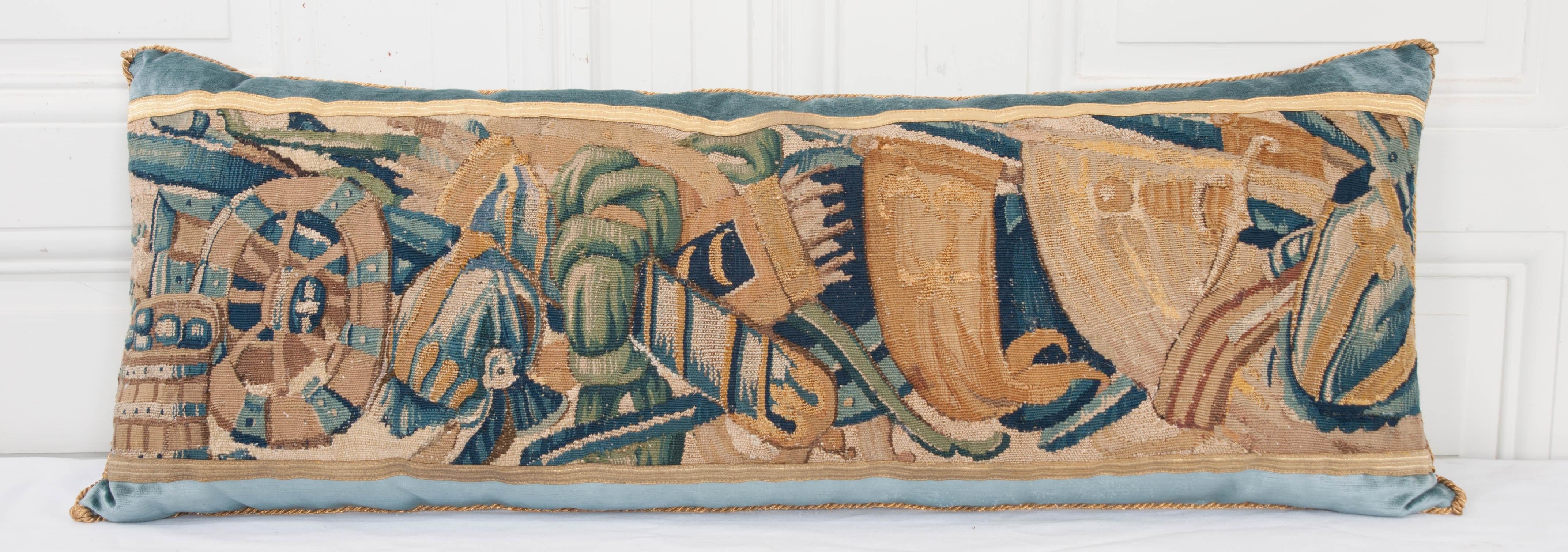 17th century tapestry fragment depicting a soldier’s gear in shades of blues, creams, greens and gold, bordered with antique gold metallic galon on Wedgwood blue velvet. Hand trimmed with vintage gold metallic cording, knotted in the corners. Down