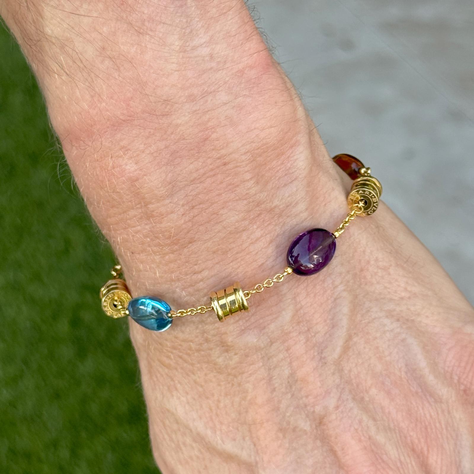 This vibrant Bvlgari B.Zero1 bracelet is crafted in luxurious 18 karat yellow gold and features the iconic tubular spiral design accented with colorful oval cabochon gemstones including garnet, blue topaz, amethyst, and citrine. The playful mix of